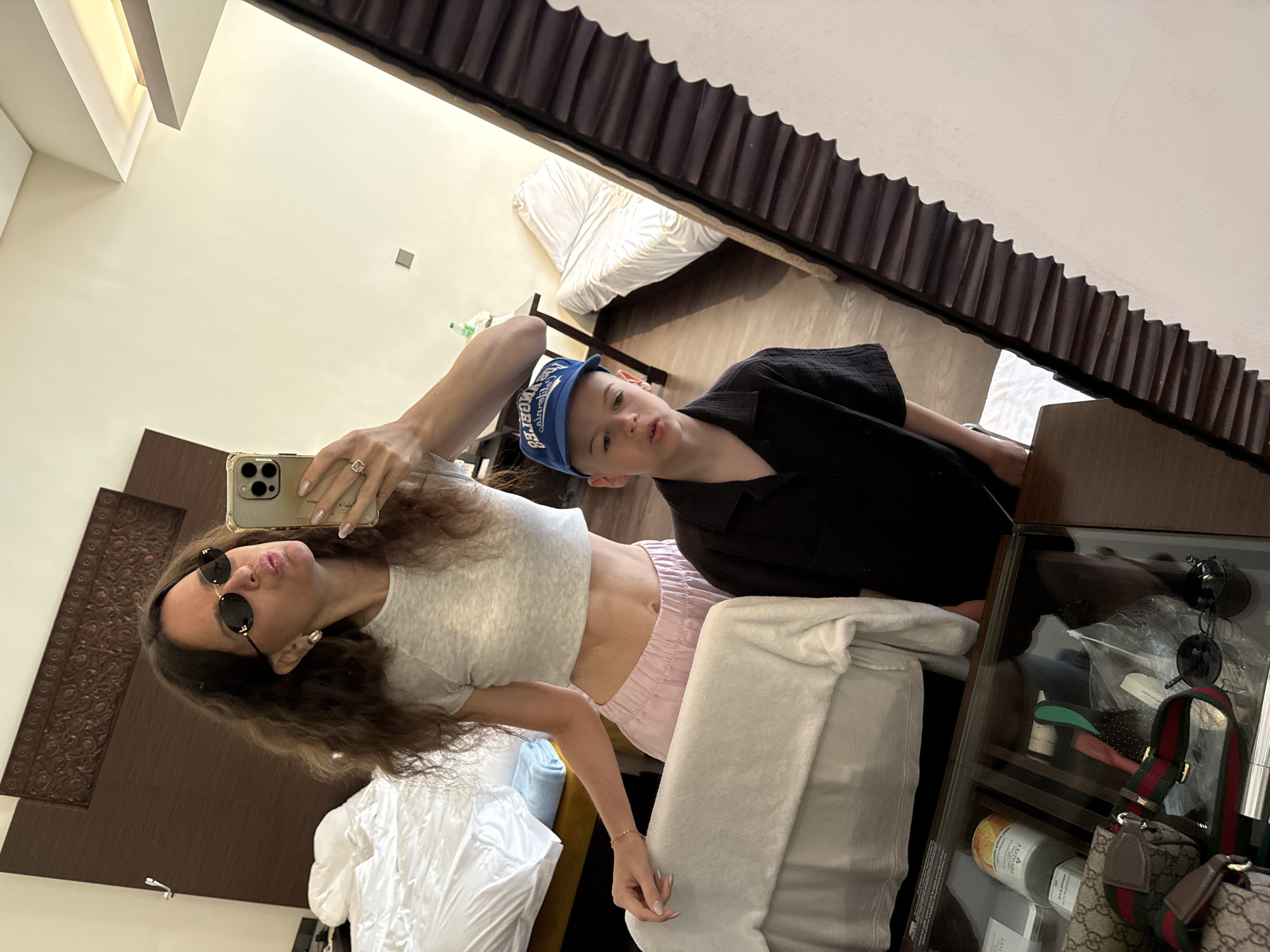 In a hotel room in Dubai, United Arab Emirates, a woman and a child stand reflected in a large mirror. The woman, with long, wavy brown hair, wears sunglasses and a light grey crop top paired with light pink pants. She holds a smartphone, angled to capture the selfie, and pouts her lips. Her left hand rests on the back of a chair, partially obscured by a white towel. The child, wearing a blue baseball cap and a black collared shirt, stands to the woman's right, looking forward with his mouth slightly open. The room is furnished with two beds with white linens, a headboard with intricate carvings, and a wooden desk with a glass top. On the desk, various items are visible, including a Gucci bag, sunglasses, and cosmetic products. The overall impression is of a casual moment captured in a hotel setting.