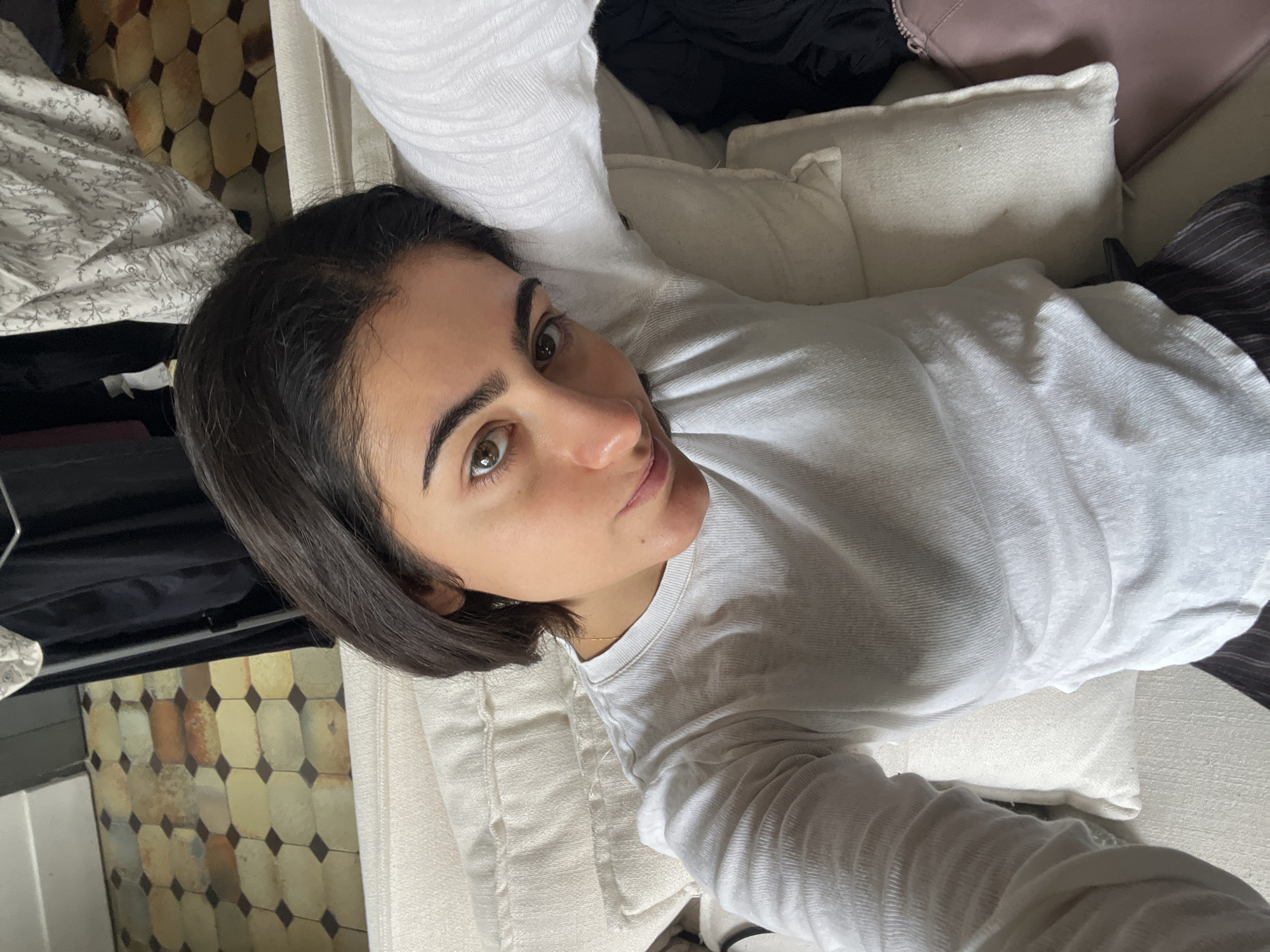 A woman with dark hair and brown eyes lies on a light-colored sofa, wearing a white long-sleeved shirt. The background includes a tiled floor, a bed with bedding, and clothing hanging on a rack. The location is possibly a residential space in Paris, France, given the architectural style and decor.
