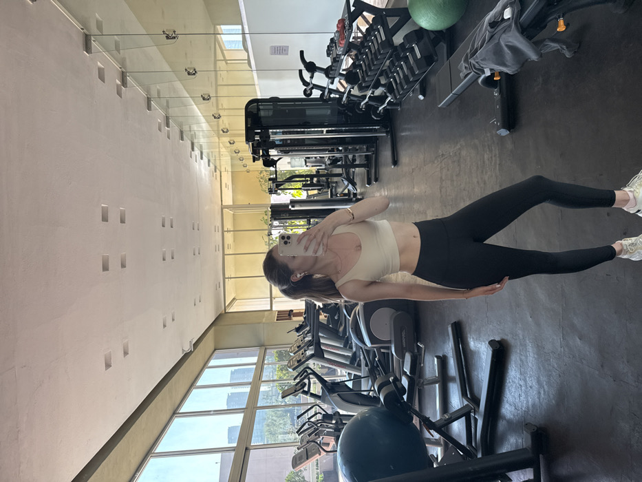 A female individual is depicted taking a mirror selfie within a gym in Mexico City, Mexico. The subject, with long brown hair in a ponytail, is wearing a light-colored sports bra, black leggings, and light-colored athletic shoes. She is holding a light-colored smartphone to obscure her face while photographing her reflection. A necklace, earrings, and a bracelet are visible accessories.
The gym features large windows on the left side, looking out onto an urban landscape with modern high-rise buildings and green areas under a clear sky. In front of these windows, multiple cardio machines, including treadmills and ellipticals, are arranged. A large blue exercise ball rests in the foreground on the left.
The right side of the gym is lined with mirrors reflecting the interior. Various weight training machines, a black dumbbell rack with multiple sets of free weights, and a black exercise bench with a grey garment draped over it are visible. A green exercise ball is on the floor near the bench. The floor is dark-colored, likely a gym-specific matting. The ceiling is light-colored and fitted with numerous square recessed light fixtures.