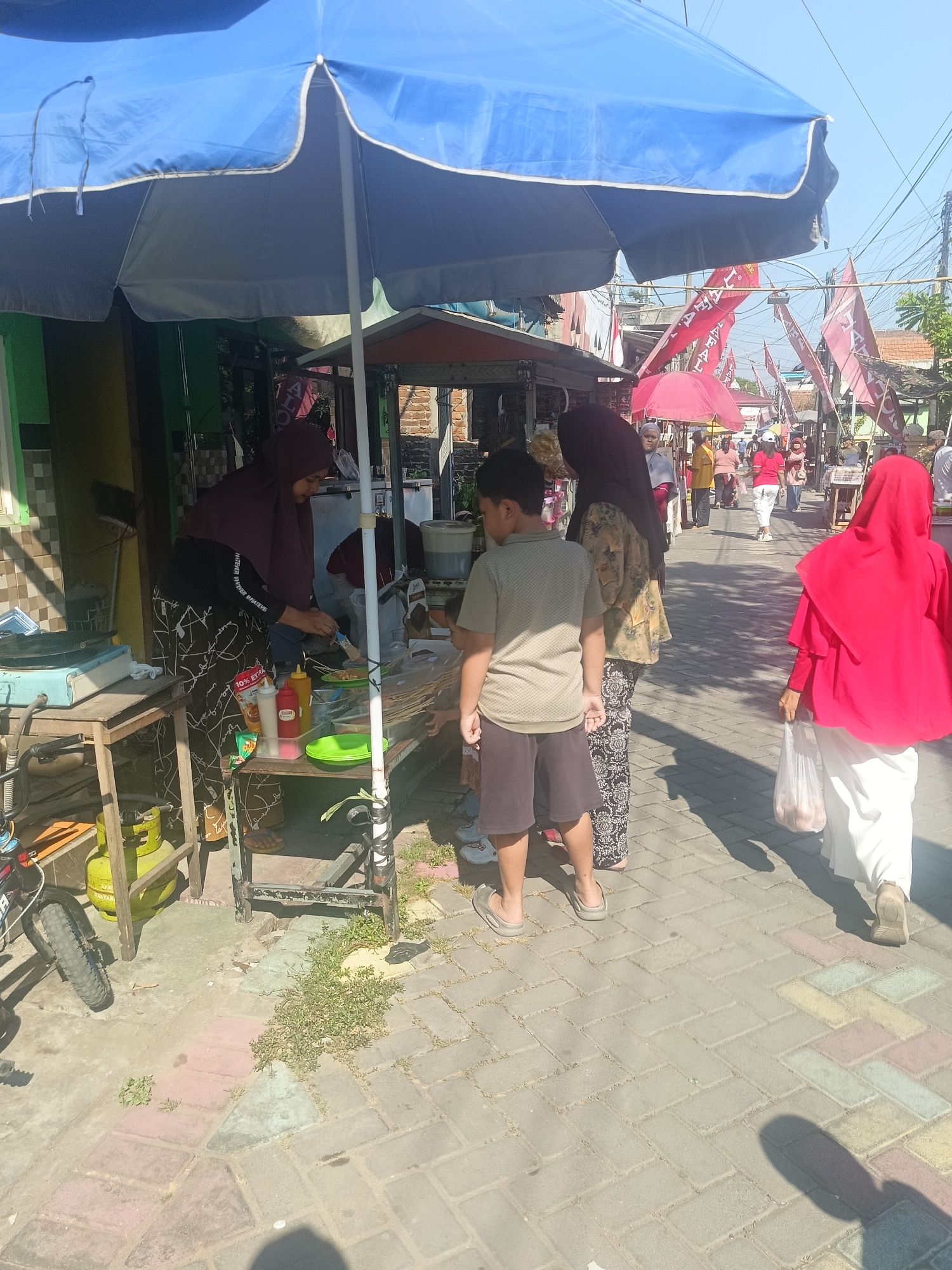 In Gedangan, Indonesia, a street scene unfolds under a large blue umbrella, suggesting an outdoor market or food stall. The ground is paved with colorful interlocking bricks. A young boy stands facing a food stall, where a woman in a maroon hijab is preparing food. She is holding a skewer and appears to be grilling or frying. Various condiments, including ketchup and mayonnaise, are visible on the stall. A green gas cylinder is positioned on a wooden table to the left of the stall. Further down the street, several people are walking, some carrying bags. Red and white flags are displayed along the street, adding a festive or patriotic atmosphere. A woman in a red hijab walks away from the camera, carrying a white plastic bag. The environment suggests a sunny day with a clear sky.