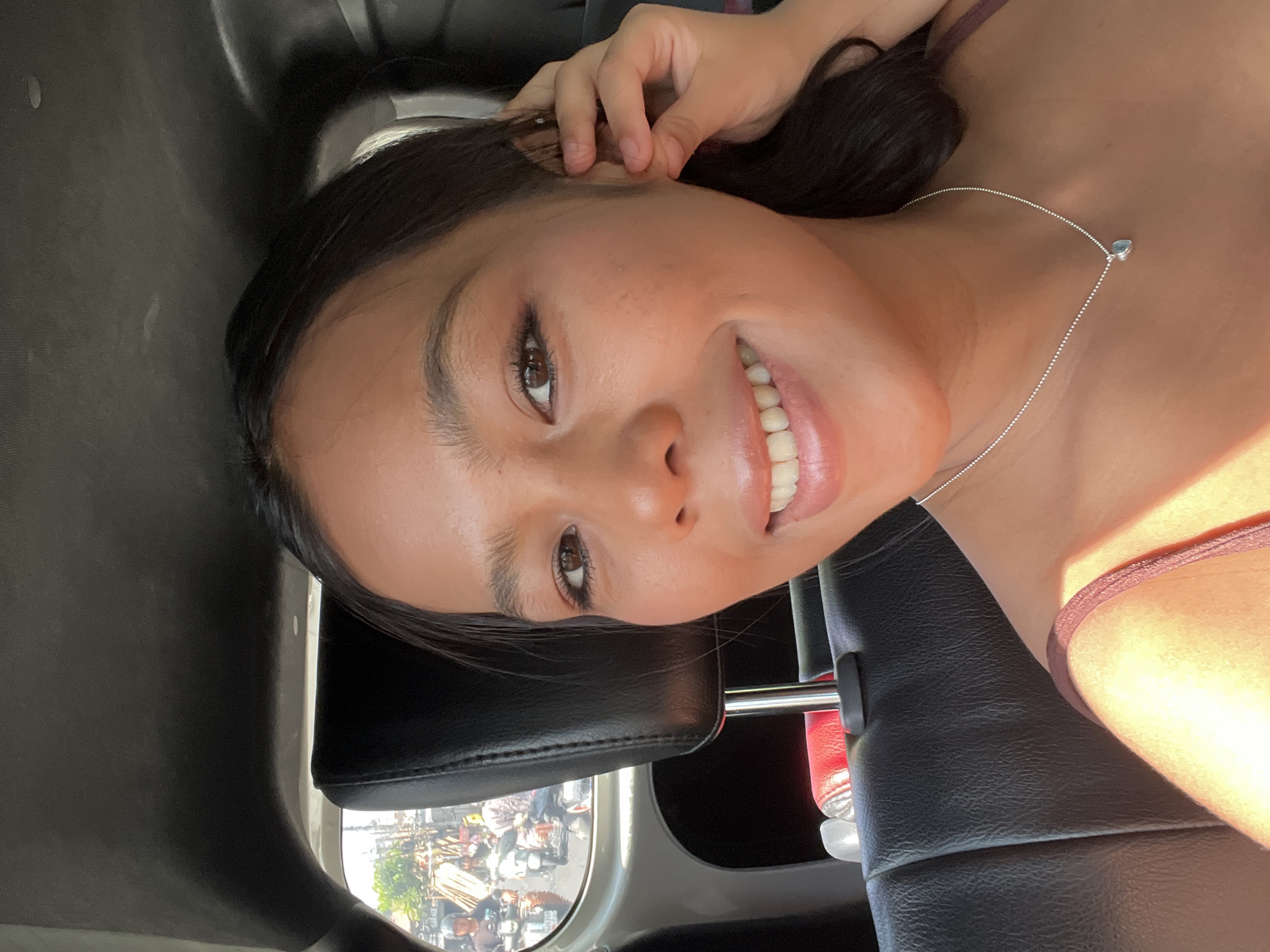 A young woman with dark hair and brown eyes is taking a selfie in the backseat of a car, likely in North Kuta, Indonesia. She is smiling and wearing a burgundy top and a silver necklace with a heart-shaped pendant. Her right hand is touching her ear. Through the car window, several motorcycles carrying passengers are visible on a street lined with trees and buildings, suggesting an urban or suburban setting. The interior of the car appears to be black leather, with a headrest visible behind the woman. The lighting suggests it is daytime, with sunlight casting shadows and highlights on her face and the car's interior.