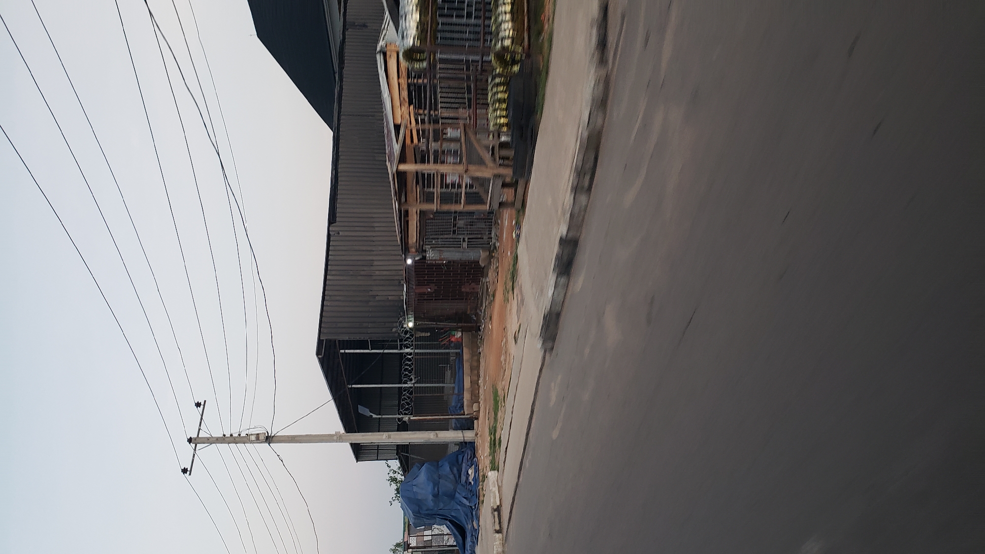 The image depicts a street scene in Jalingo, Nigeria, likely taken from a moving vehicle due to motion blur. In the foreground, a paved road with a distinct curb runs diagonally across the frame. To the left of the road, a section of sidewalk is visible. Beyond the sidewalk, an area of uneven, sandy ground is present, littered with debris.
Dominating the midground are several structures. On the left, a large, dark blue tarpaulin is draped over an unseen object. Adjacent to this is a large, corrugated metal building with an open-fronted section revealing metal framing and what appears to be barbed wire fencing. To the right of this building are several smaller, enclosed structures, some with metal bars and wooden elements. One of these appears to be a small wooden kiosk or shelter. In the background, a dark, sloped roof structure is visible. Overhead, a utility pole with multiple wires stretches across the sky, which is a pale, overcast gray. The overall impression is one of an industrial or storage area bordering a street. No people are visible in the frame.