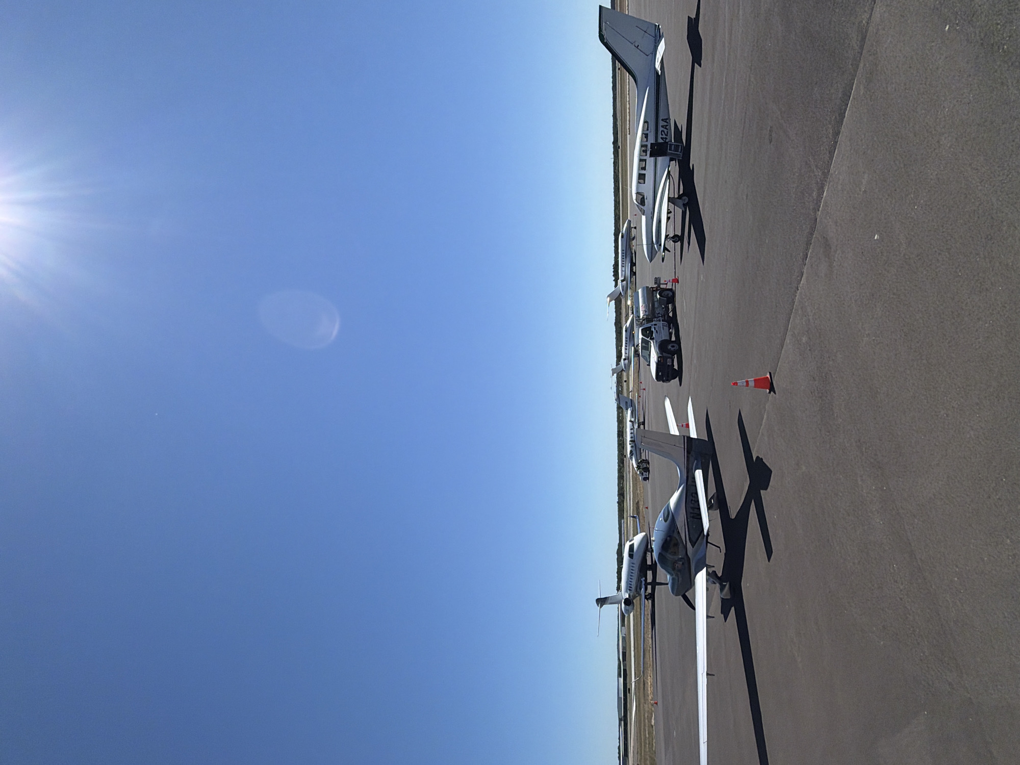 This is a wide-angle, eye-level, outdoor, daylight, full shot of an airport tarmac in the Town of Southampton, United States of America. The sky is a clear, bright blue, with the sun visible in the upper right corner, creating a lens flare. The tarmac is asphalt. In the foreground, a small, single-engine airplane with a silver fuselage and white wings is parked, facing right. Its registration number appears to be N132JW. To the right of this airplane, a larger, twin-engine propeller aircraft with a white fuselage and dark green trim is parked, facing right. Its registration number is 42AA. Behind these two planes, further down the tarmac, are several other aircraft, including a white jet airplane with a dark stripe on its tail. A small white truck with a fuel tanker trailer is parked to the left of the twin-engine propeller aircraft. Several orange traffic cones are placed on the tarmac around the parked vehicles and aircraft. The background consists of a flat, treeless landscape. There are no people visible in the image.