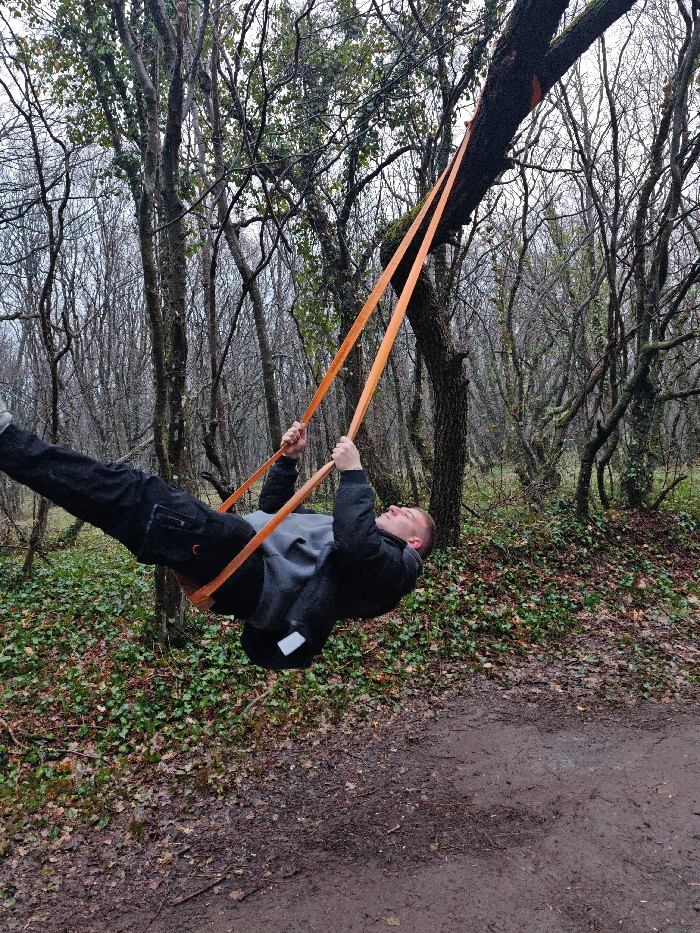 An adult male subject is depicted suspended horizontally in a wooded area, likely within or on the outskirts of Shumen, Bulgaria. The subject, wearing a dark jacket, a grey hooded sweatshirt, and dark trousers, is holding two orange fabric straps with both hands. These straps are secured over a moss-covered, horizontal tree branch originating from a tree on the right side of the frame. The subject's back is oriented towards the ground, with his head slightly raised and legs extended diagonally upwards to the left. The ground is a mixture of mud, fallen leaves, and patches of green ivy-like ground cover. Numerous deciduous trees, largely bare of foliage, are visible throughout the background, indicative of a late autumn or winter season under overcast skies. A dirt path runs along the lower right portion of the image.