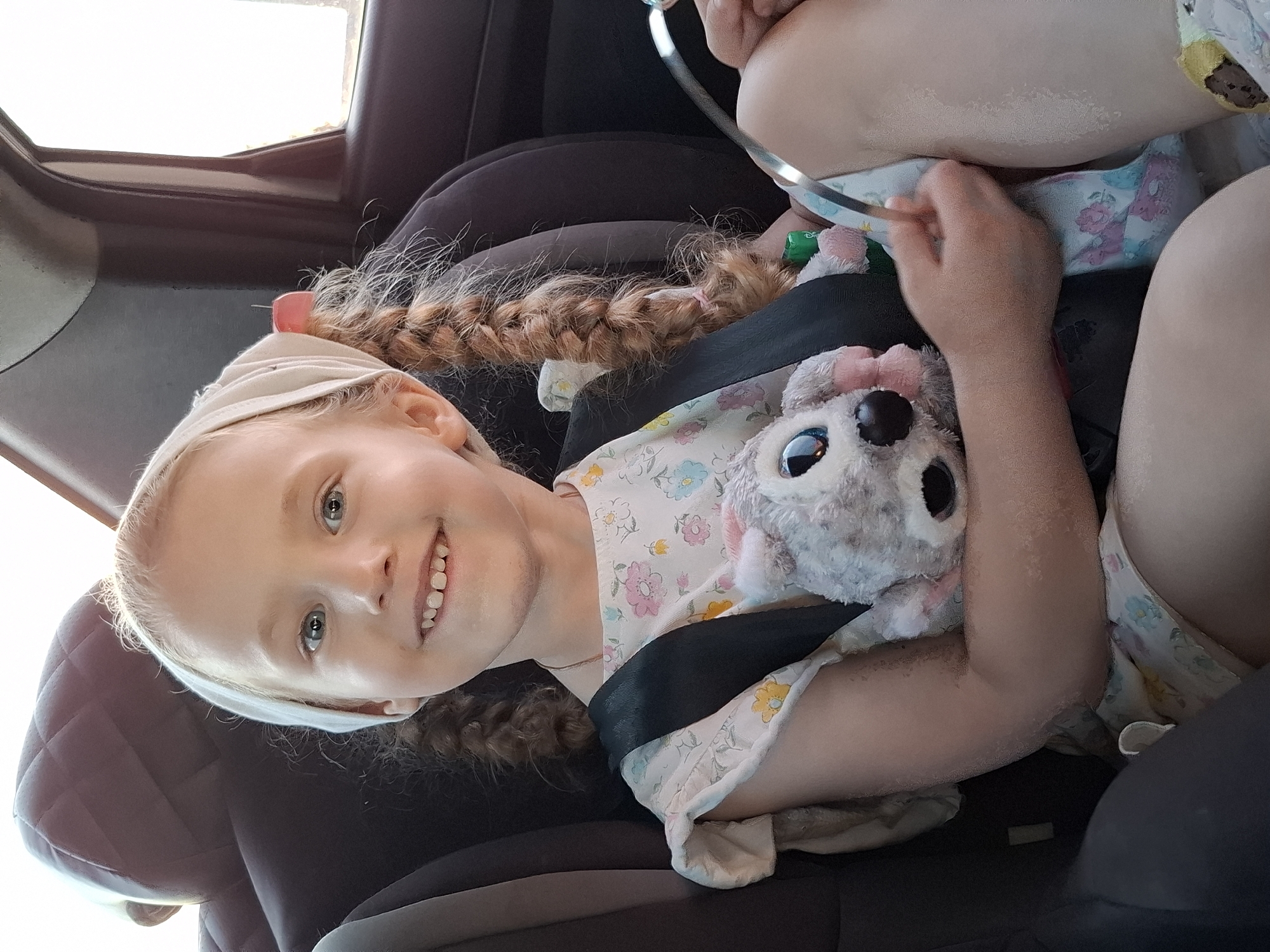 A young blonde girl with braided hair and a light-colored headband is sitting in a car seat. She is wearing a floral-patterned dress and secured with a black seatbelt. She is holding a gray stuffed animal with large eyes. The interior of the car is visible, with a window and dark-colored upholstery. The image does not provide any information about the location in Гаревая, Russia.