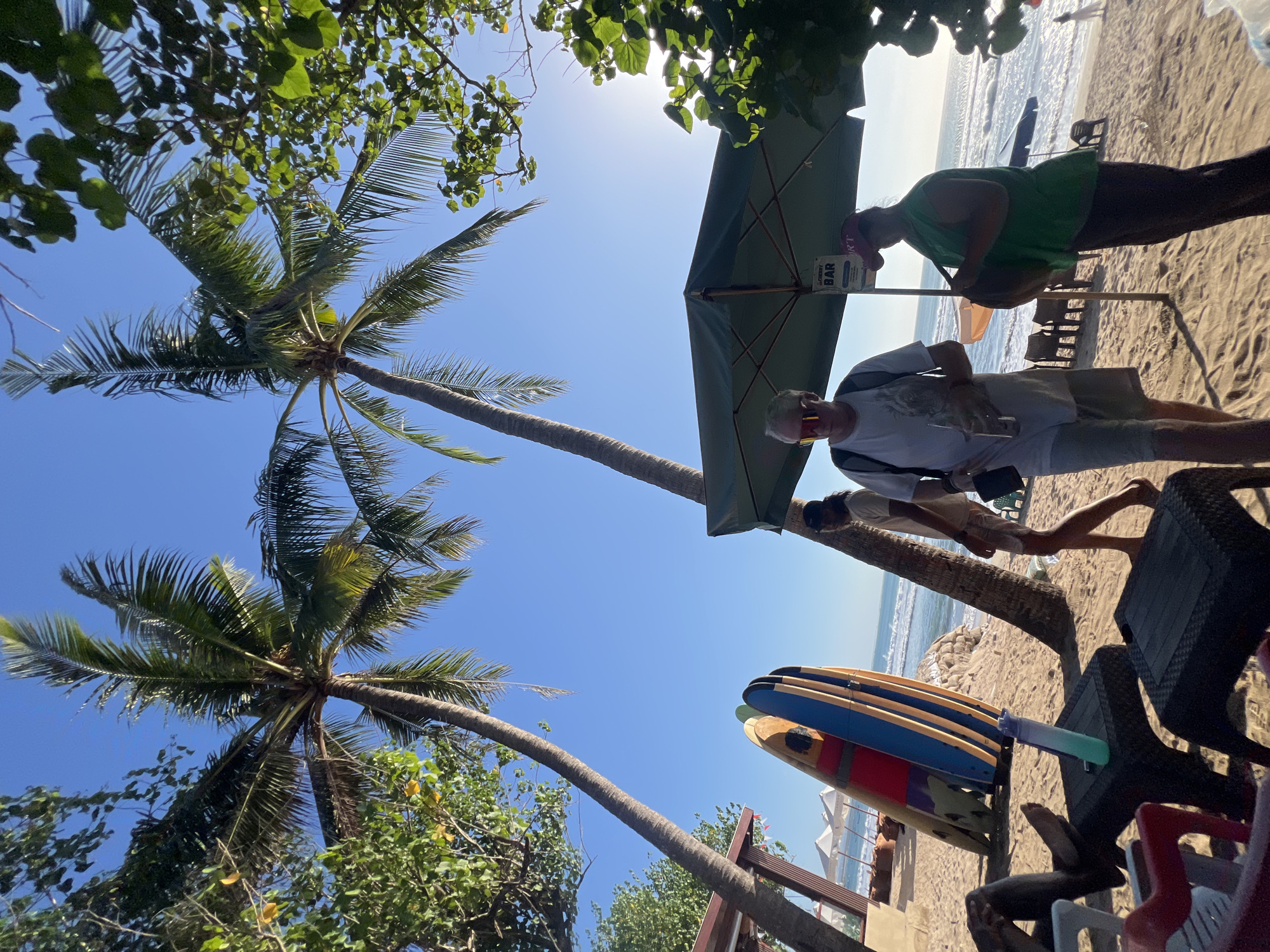 A low-angle shot captures a sunlit beach scene in Kuta, Indonesia, dominated by two tall palm trees against a clear blue sky. In the foreground, on the sandy beach, several surfboards are propped upright. Two people are walking near a large green beach umbrella. One, a man with sunglasses and a backpack, is holding a drink. The other, a woman wearing a green shirt and a red cap, is carrying a large bag. Other people are visible in the background, along with beach chairs, umbrellas, and the ocean with gentle waves. The overall impression is a relaxed, tropical beach atmosphere.