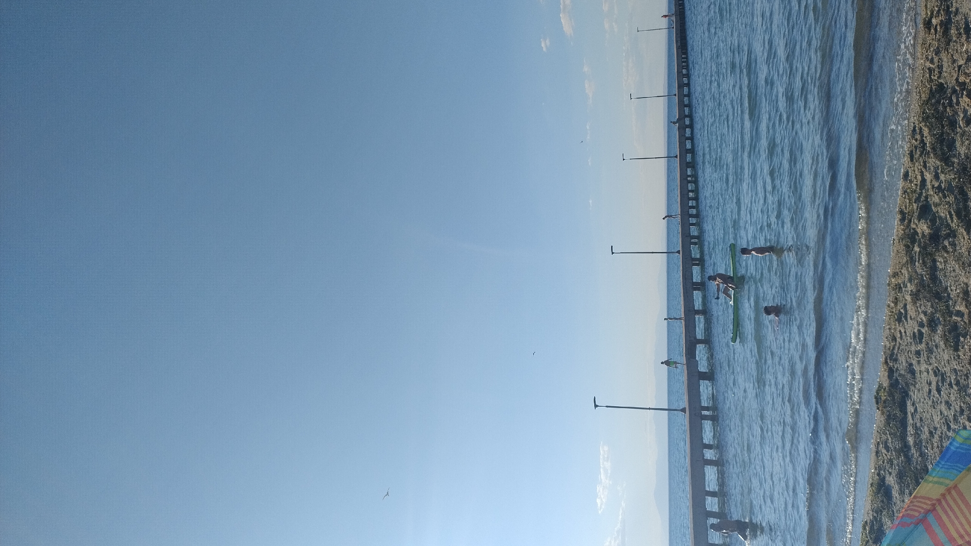 In Perea, Greece, the image captures a serene coastal scene under a clear blue sky. A long pier, lined with lampposts, stretches across the horizon. The sea is calm, with gentle waves lapping the sandy shore in the foreground. Several individuals are enjoying the water and the pier. On the pier, people are seen walking and standing. In the water, one person is paddleboarding on a green board, while others are swimming. The water appears to be shallow near the shore. In the distance, hazy mountains are visible, adding depth to the landscape. A colorful beach towel is partially visible in the lower left corner of the frame. The overall atmosphere is one of leisure and relaxation on a sunny day.