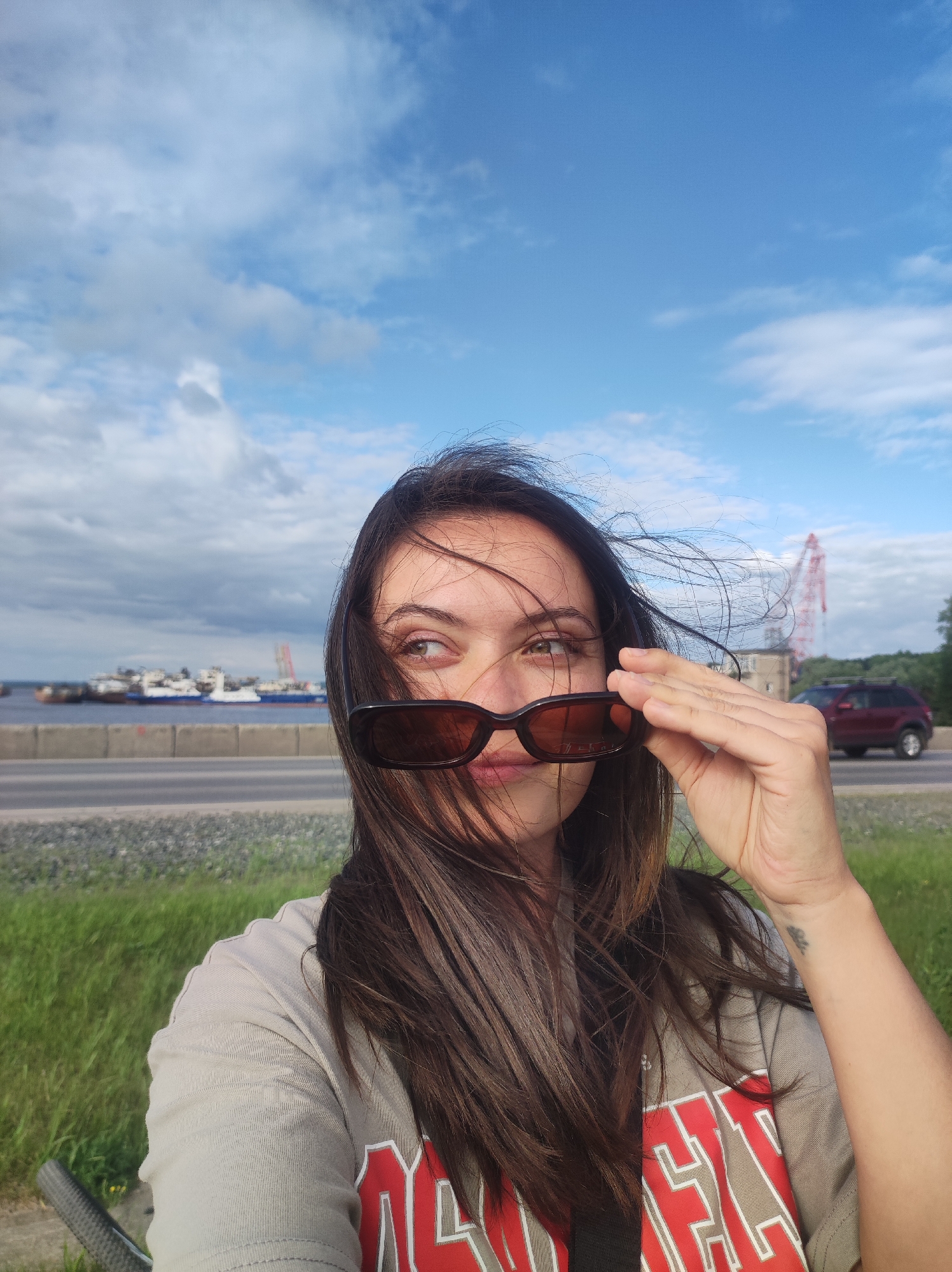 A medium shot of a young woman looking to her right, holding her sunglasses up to her eyes with her right hand. She has long, dark brown hair that is blowing across her face. She is wearing a light tan t-shirt with red and white lettering on it. She is outdoors with a blue sky with white clouds in the background. In the middle ground, there is a body of water with several boats, a highway with a red car driving on it, and a red crane. The foreground consists of green grass and a bicycle handlebar. The location is outdoors in Unknown, Unknown.