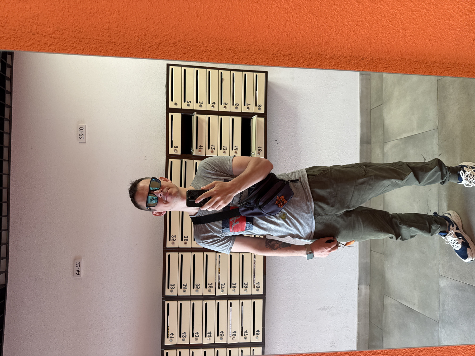 A man stands in a hallway near a wall with a bank of mailboxes, as reflected in a mirror. The man is wearing sunglasses, a gray t-shirt, olive-green pants, and two-tone sneakers. He's holding a phone in his hand and wearing a small messenger bag across his chest. The mirror shows a close-up reflection of the man and a view of the mailbox wall. The mailboxes are labeled with numbers. Above the wall, the mirror reflects an orange ceiling or wall. Small labels on the wall read "S2-11" and "S3-14." The hallway floor is tiled. The image was taken in Krasnodar, Russia.