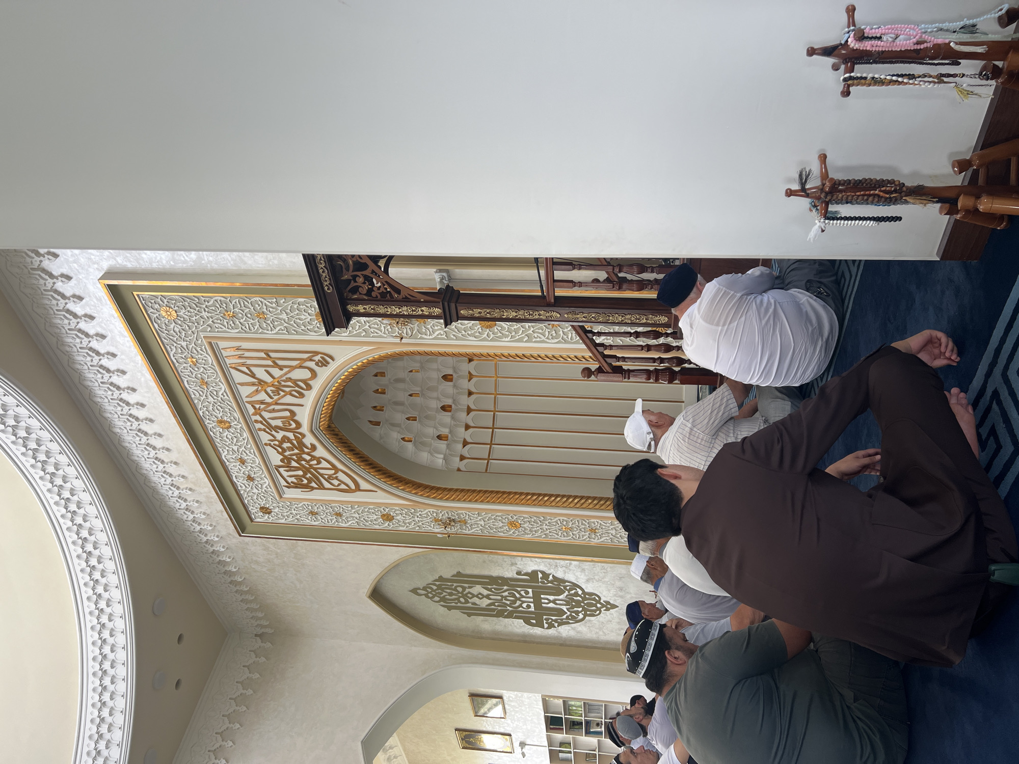 The image depicts the interior of a mosque in Karamanas, Uzbekistan, during prayer. A group of men, dressed in traditional Islamic attire and caps, are seated on a blue carpeted floor in a prayer posture, facing the mihrab, which is adorned with ornate Arabic calligraphy and golden floral motifs. The walls of the mosque are decorated with intricate patterns, and a wooden minbar stands to the right of the mihrab. Prayer beads are displayed on wooden stands near the wall. The lighting is soft and natural, suggesting it might be daytime.