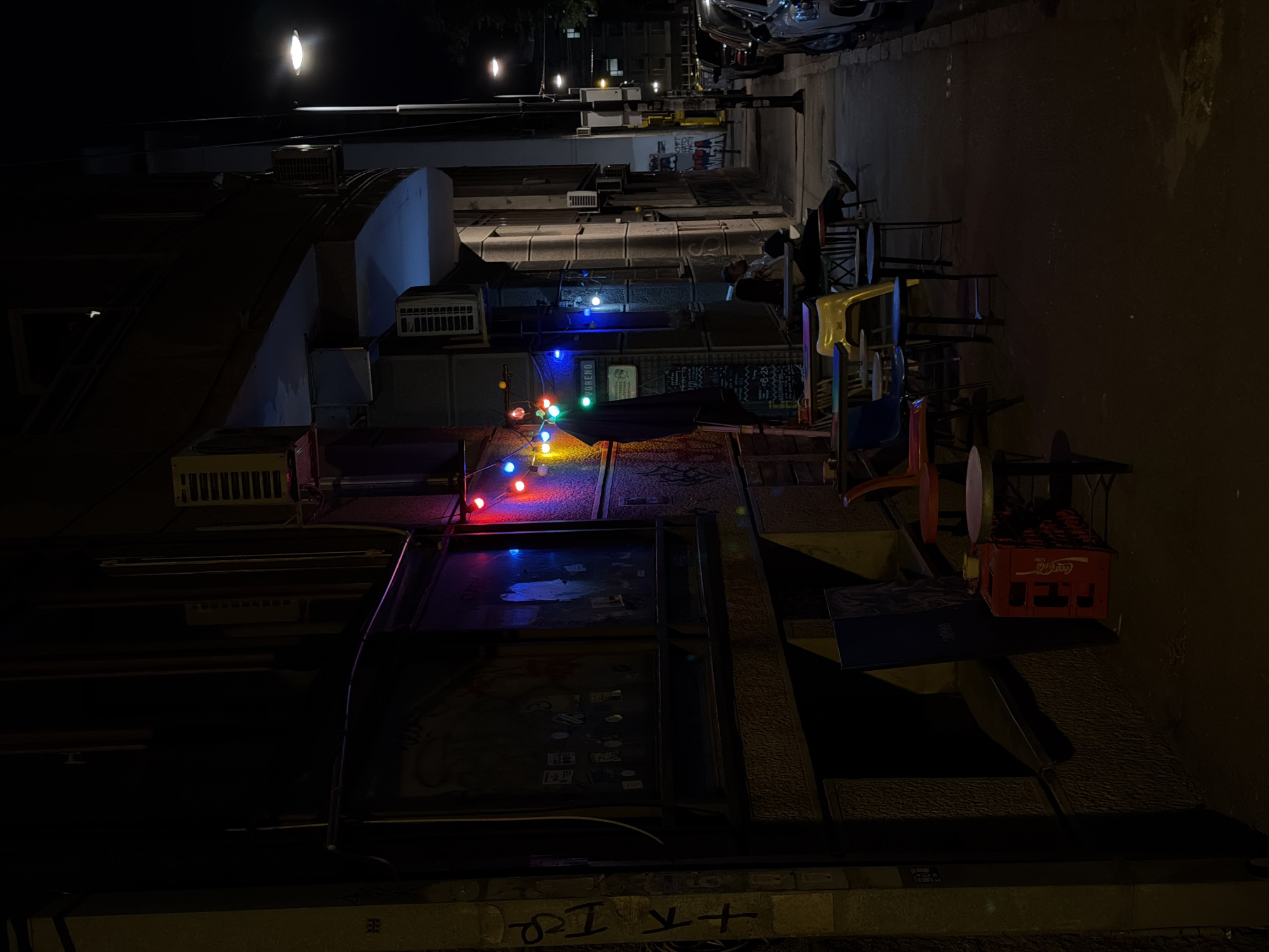 The image shows an alleyway at night, likely in Vracar Urban Municipality, Serbia. The alley is dimly lit, with a string of colored lights strung across the buildings. The buildings appear to be older, with exposed air conditioning units and other infrastructure. There are tables and chairs set up outside, suggesting a cafe or restaurant. A red Coca-Cola crate is also visible. Graffiti can be seen on the walls. There are several stacked chairs.