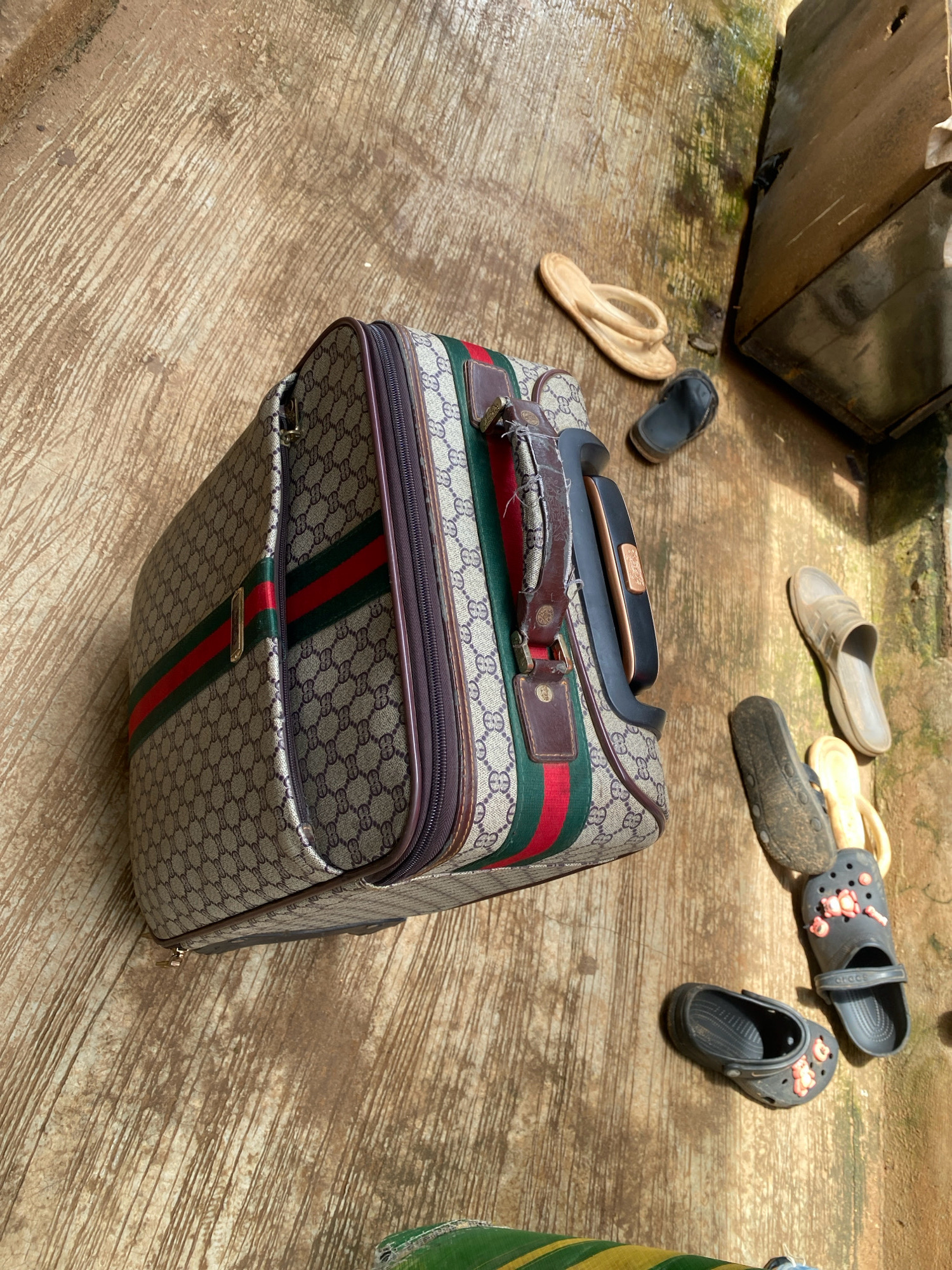 A Gucci-patterned suitcase lies on a wet, concrete surface in Iworoko, Nigeria. Scattered around the suitcase are several pairs of footwear, including flip-flops, slides, and Crocs. To the right of the suitcase, a wooden structure is visible, partially obscured by shadows. The ground appears weathered and stained, with visible water puddles reflecting the environment. The lighting suggests an outdoor setting, likely during daylight hours.