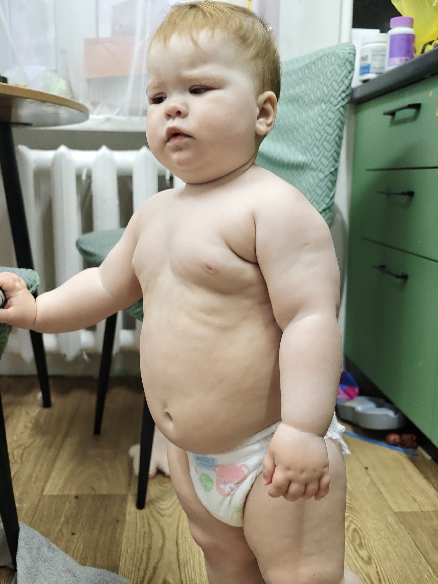 A toddler stands indoors, wearing a diaper. The background includes a radiator, a table, a chair with a green patterned seat, and green cabinets. The location is Chaikovsky, Russia.