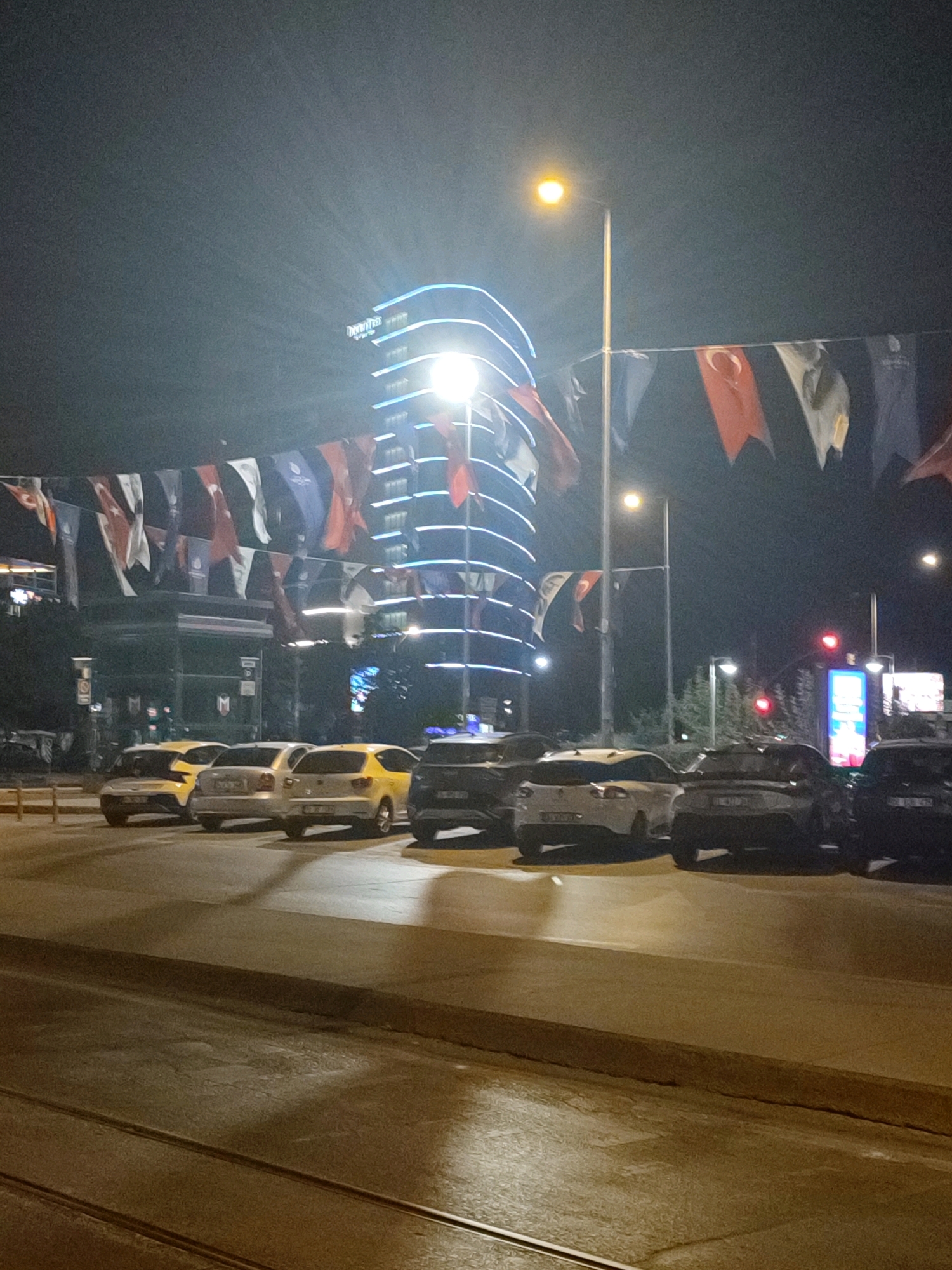 The image depicts a nighttime street scene in Kadıköy, Turkey. A modern, tall building with blue, illuminated trim stands prominently in the background, displaying the text "Divan" near its top. In the foreground, several cars are parked along the street, including two yellow taxis and darker sedans. Decorative flags, some featuring the Turkish flag, are strung across the street, adding a festive or commemorative element to the scene. Streetlights illuminate the area, casting a warm glow on the wet pavement and car surfaces. Tram tracks are visible in the lower part of the image, indicating the presence of a tram system. The overall atmosphere is that of a well-lit urban environment at night.
