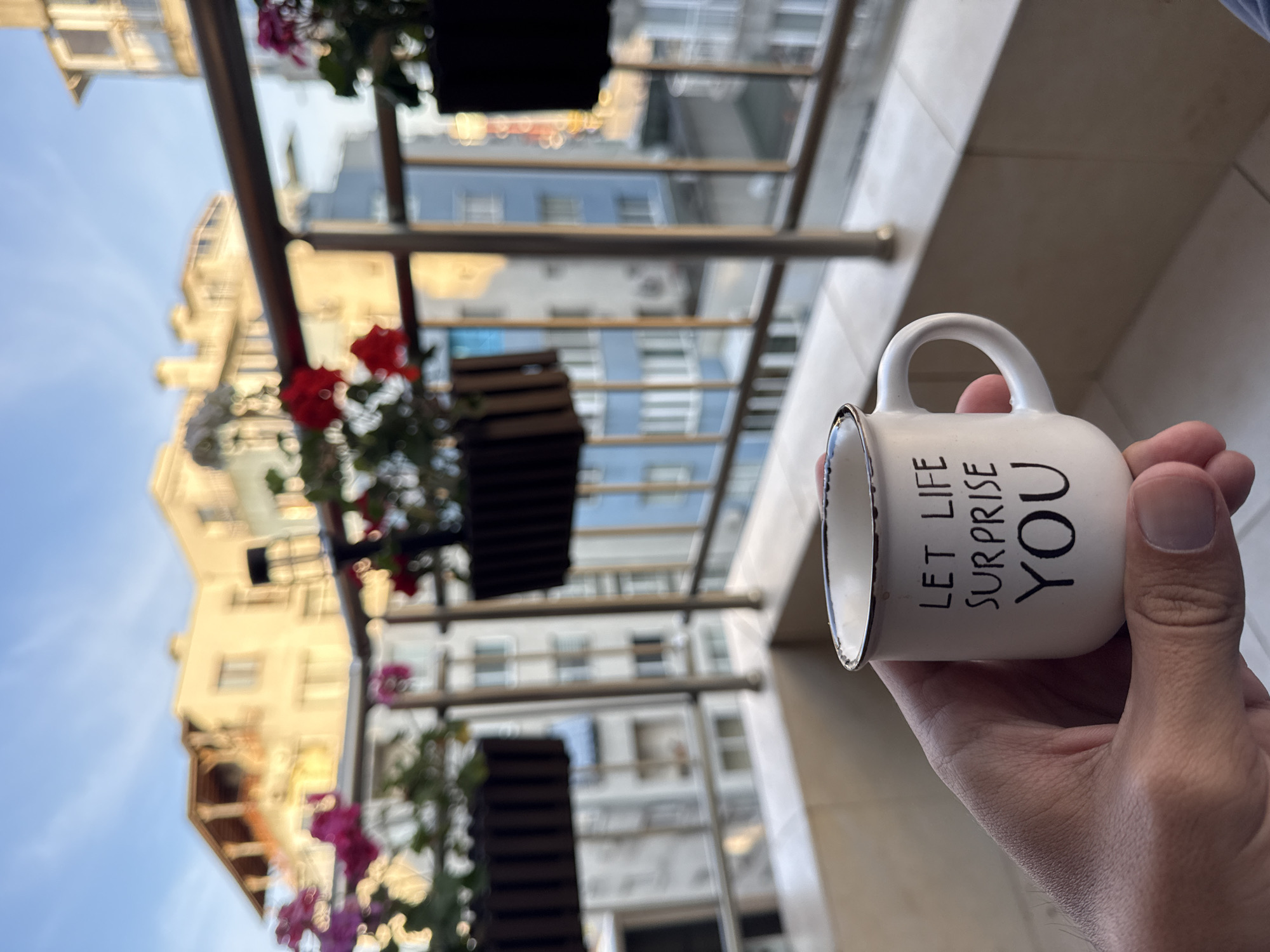A person's hand holds a white mug with the words "LET LIFE SURPRISE YOU" in black lettering. The background is a blurred urban scene, suggestive of the city of Burgas, Bulgaria, featuring buildings of varying colors, from light tan to blue-grey, under a blue sky. A metal balcony railing with flower boxes is visible in the foreground.
