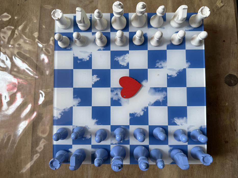 The scene, located in Carnin, France, depicts a chessboard resting on a light-colored wooden surface. The chessboard features a blue and white grid, with the blue squares displaying white cloud patterns. Thirty-two chess pieces are arranged in their standard starting positions. The pieces on the left side are light blue, consisting of two rooks, two knights, two bishops, one queen, one king, and eight pawns. The pieces on the right side are off-white, similarly consisting of two rooks, two knights, two bishops, one queen, one king, and eight pawns. A small, flat, red heart-shaped object with a visible hole near its top left edge is placed on a central blue square of the board. Distorted, wavy reflections, incorporating bright light and red tones, are visible on the wooden surface above the chessboard.
