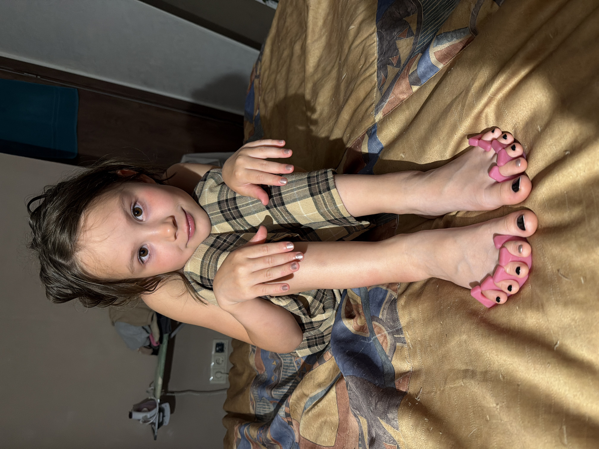 A young girl with dark hair and brown eyes lies on a bed with a patterned comforter. She is wearing a plaid dress. Her feet are extended, and she has toe separators on her right foot. The toe separators are pink and are positioned between her toes, which are painted with black and silver nail polish. Her hands are also visible, with her nails painted the same colors. The lighting in the room suggests it is daytime, with shadows cast across the comforter. The background shows a wall with a light switch and an electrical outlet, as well as some blurred furniture. The overall impression is of a casual, indoor scene. The location is identified as Perm, Russia.