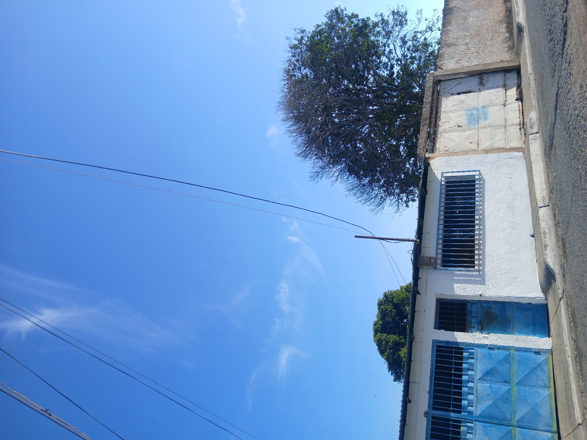 The image shows a building with a partially obscured facade. The building is white with blue accents on the doors and windows, and it appears to have metal bars on the windows. A tree is visible near the building. Power lines cross the sky. The sky is blue with some light clouds. The location is Porlamar, Venezuela.