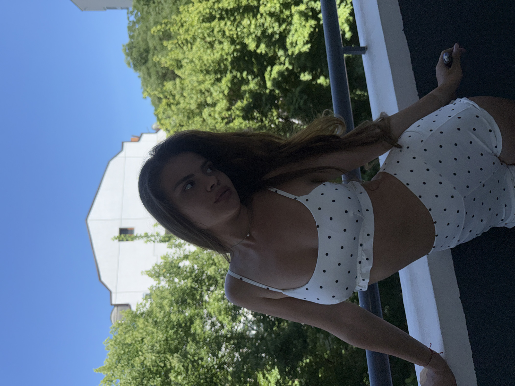 A young woman with long, brown hair, wearing a white polka-dot crop top and matching shorts, stands on a balcony. She is leaning on the railing, with buildings and lush green trees in the background under a clear blue sky. The scene appears to be set in the city of Plovdiv, Bulgaria, possibly in a residential area with apartment buildings. The woman's gaze is directed to the side, and she holds a small object, possibly a cigarette lighter, in her right hand.