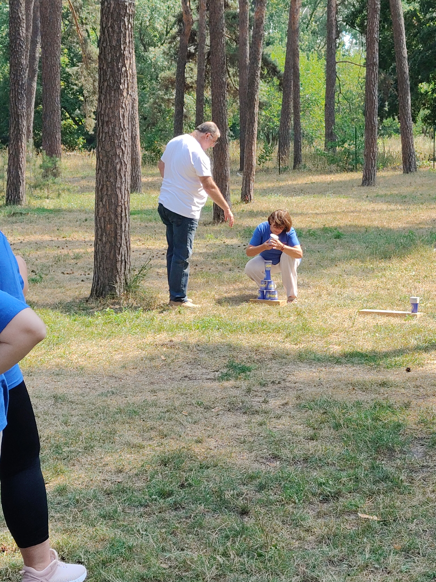 In Cherkasy, Ukraine, in a wooded area with dry grass and scattered pine trees, two individuals are engaged in an outdoor activity. A man, wearing a white t-shirt and blue jeans, stands in the foreground, his left arm extended. Crouching in the mid-ground is a person in a blue t-shirt and off-white pants, focused on a stack of blue cans. To the right, a wooden plank with a blue can rests on it. The scene appears to be a recreational area, possibly a park or campground, under natural daylight.