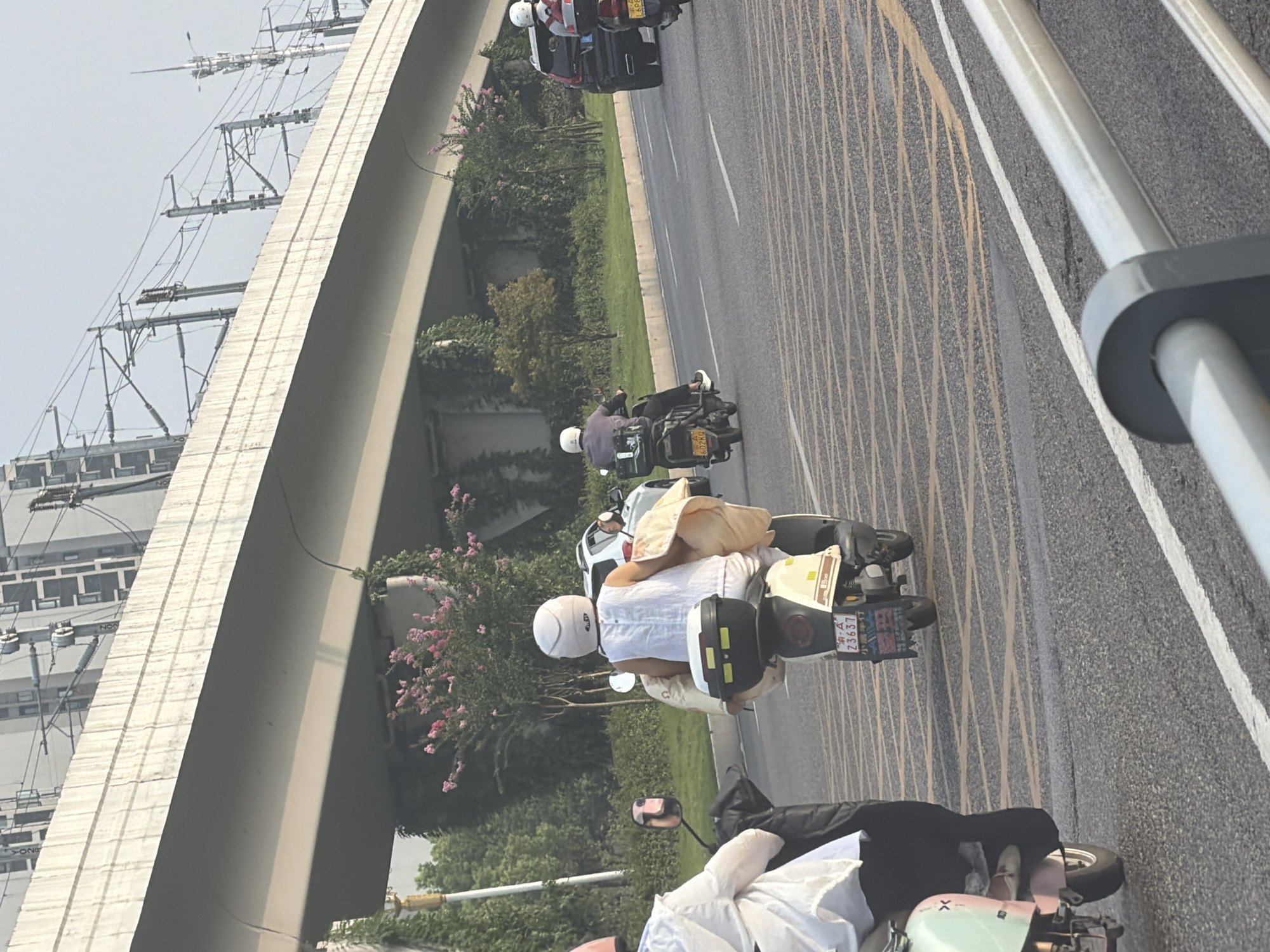 The image depicts a street scene in Jiangjin District, Chongqing, China, featuring multiple individuals on motorcycles and a car. In the foreground, a person rides a light-colored electric scooter. To their right and slightly behind, a person in a white outfit and helmet rides another electric scooter, carrying a yellow blanket. Further back, another individual on a motorcycle wearing a helmet is visible. A dark SUV is present in the right lane, and a black motorcycle is further ahead. The road is marked with yellow and white lines. An elevated structure, likely an overpass or railway bridge, looms above, with visible power lines and poles. Green trees and bushes line the sides of the road. The sky is overcast.