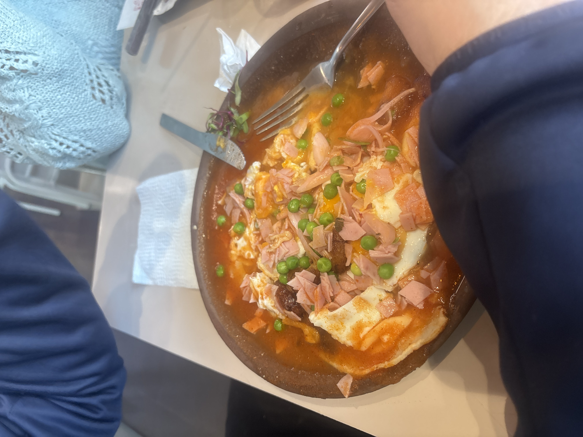 The image shows a dish of Huevos a la Poblana (Puebla-style eggs) being eaten. The dish, served in a dark brown clay bowl, contains eggs fried in a tomato-based sauce, topped with diced ham and green peas. A fork and a knife are positioned on the rim of the bowl. A person wearing a blue sweater is seated at the table, and another person wearing a blue crocheted sweater is visible in the left corner. The dish is on a light-colored table at a restaurant, presumably in the city of Puebla, Mexico.