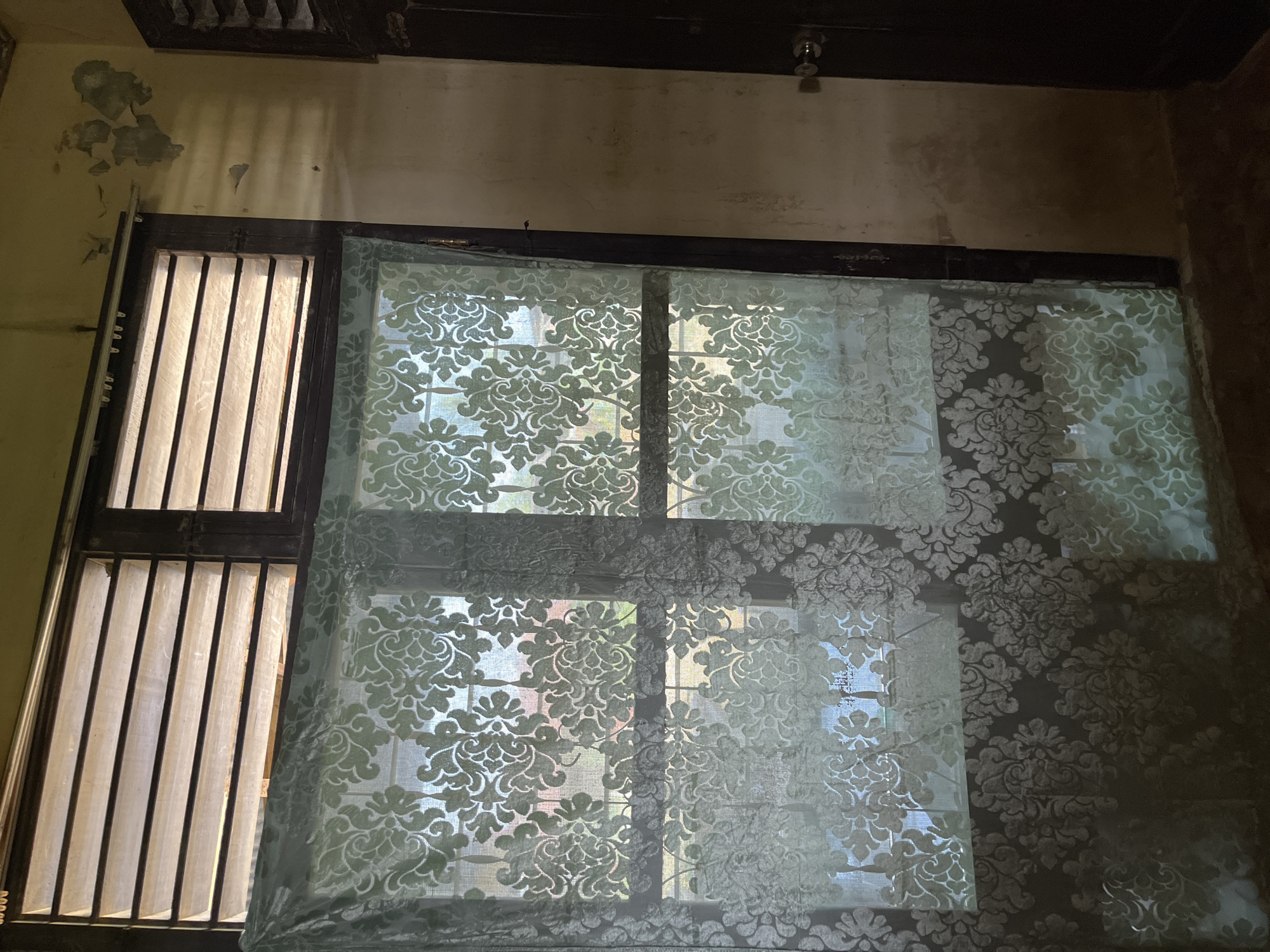 This is an indoor photograph, likely taken within a residential building in Kota Kuala Simpang, Indonesia. The central focus is a window covered by a sheer, light green curtain with an ornate floral pattern. The window itself appears to be a louvre window, with adjustable slats allowing for ventilation. The curtain is draped across the lower portion of the window, obscuring the view outside but allowing light to filter through.
To the right of the window, a dark-colored door is visible, featuring a circular silver doorknob. The wall adjacent to the window and door exhibits signs of wear and tear, with patches of peeling paint in a lighter shade, suggesting an aged interior. The overall lighting in the room is dim, with light emanating from the window, casting shadows on the wall. The floor is not clearly visible, but appears to be a darker, possibly concrete surface. There are no people or animals present in the image.
