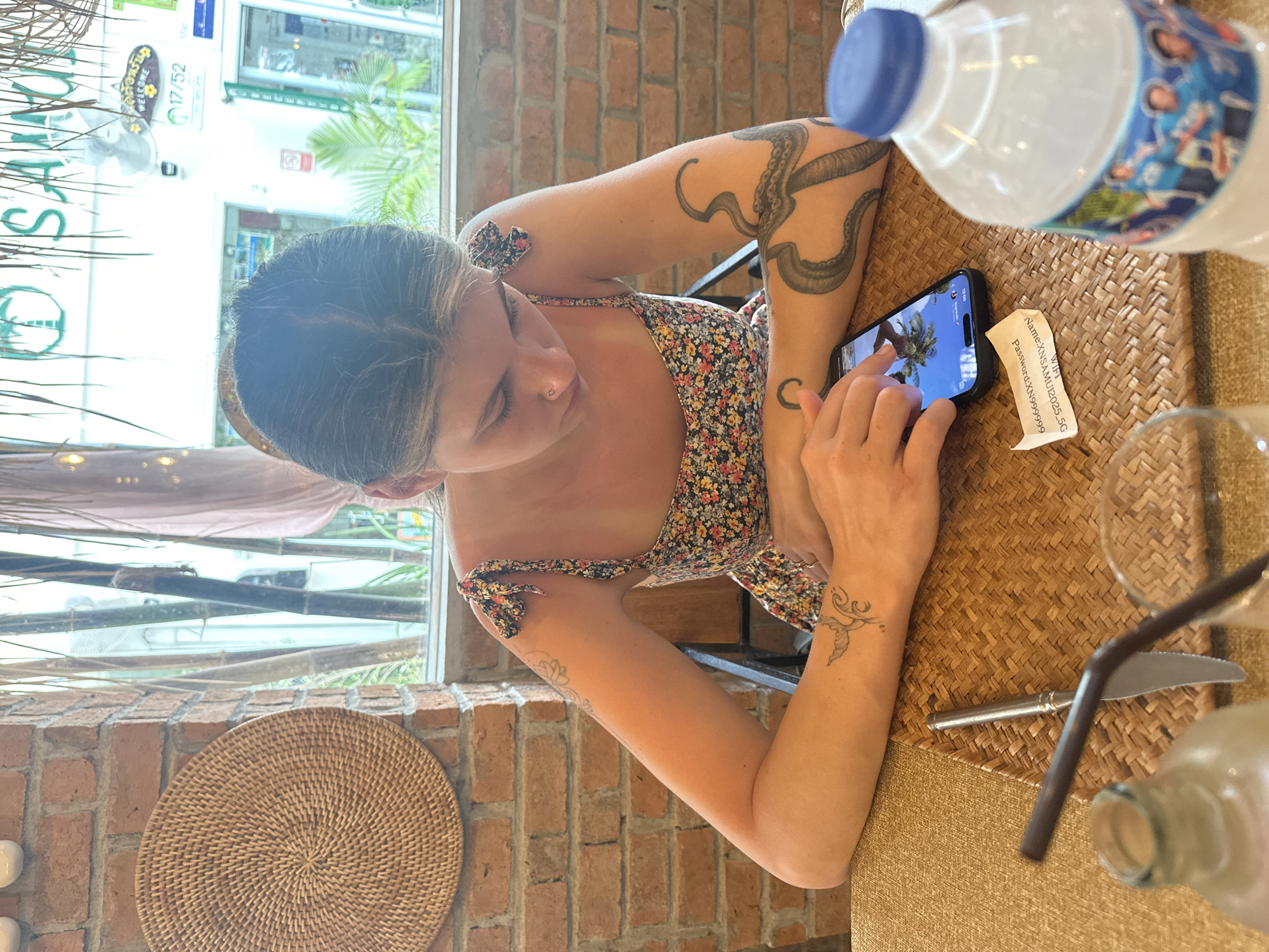 A woman with an octopus tattoo on her left arm is at a table with a woven placemat in a cafe. She is looking at a smartphone in her right hand. A water bottle, a paper with WIFI information, a glass, a straw, a knife, and a bottle are on the table. The cafe has a brick wall and a window overlooking the street. The cafe is in Baan Chaweng, Thailand, as indicated by signage outside.