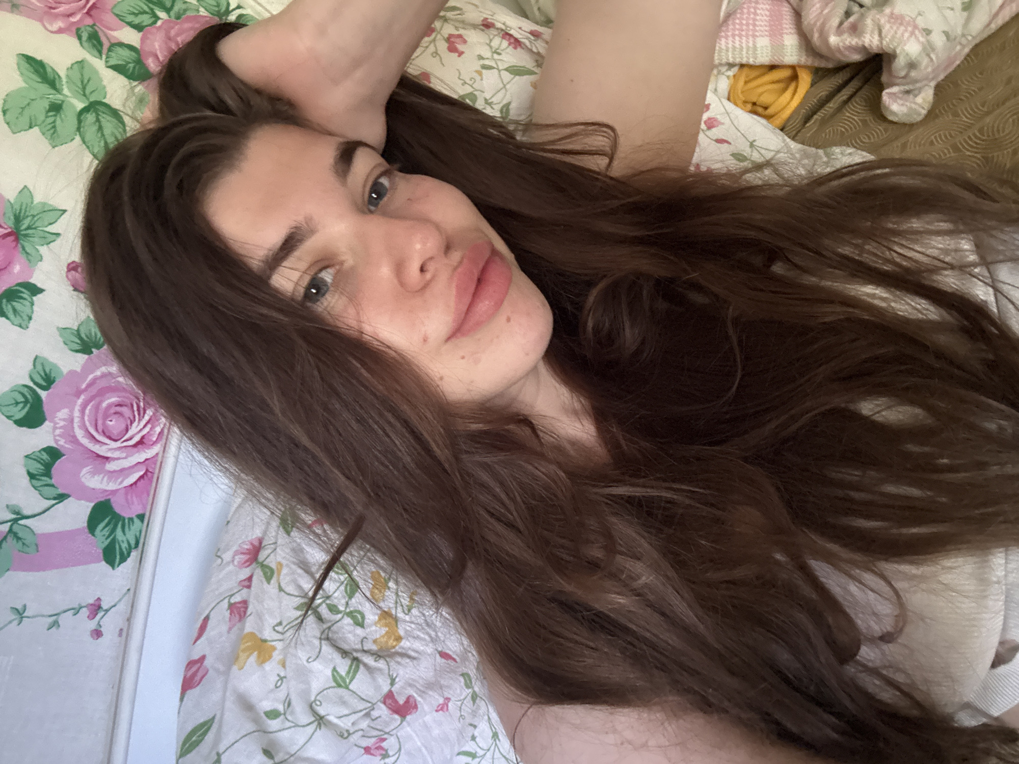 A young woman with long, dark brown hair is lying in bed, with her head resting on her hand. She is wearing a white top. The background is a floral patterned duvet and pillows. The scene is indoors, likely a bedroom. There is no indication of the specific location within the city of Мещёры, Russia.