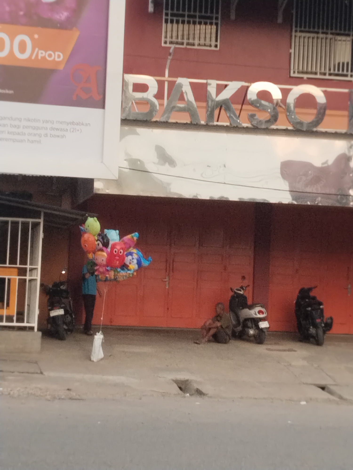 The image captures a street scene in Lhokseumawe, Indonesia, with a building facade prominently featuring a metallic sign that reads "BAKSO". In front of the building, a person is standing and holding a large bunch of colorful cartoon-themed balloons. To the left of the balloons, a motorcycle is parked. Further to the right, another motorcycle is parked and a person is sitting on the ground next to it, leaning against a dark-colored motorcycle. The street appears to be paved, with a concrete sidewalk and a drainage grate visible. The building itself has an orange-red wall and windows with metal bars. The overall lighting suggests it is daytime.