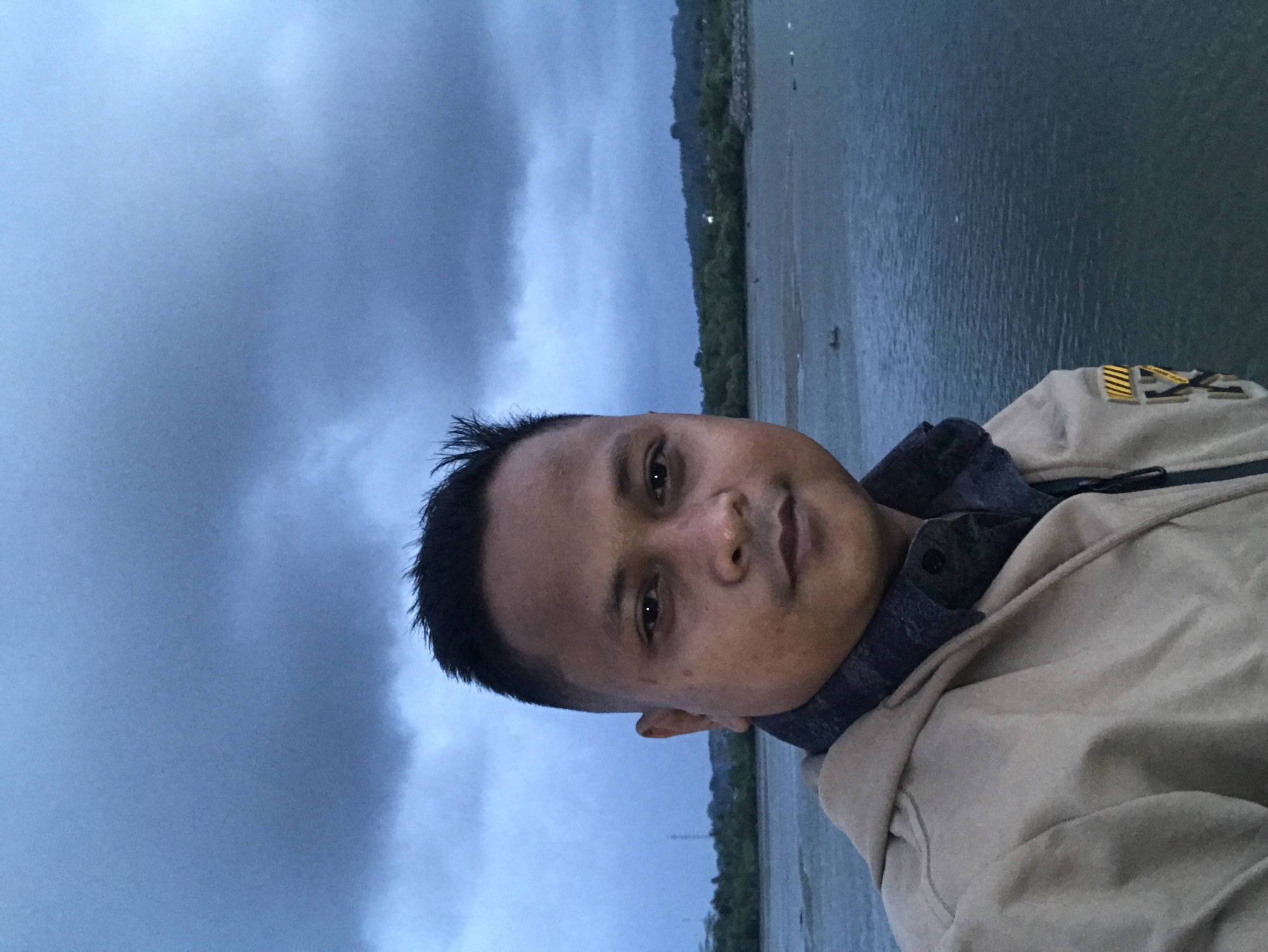 The image is a selfie of a man with short, dark hair and light brown skin. He is wearing a dark blue shirt and a light brown jacket. He is likely standing near the coast in Lhokseumawe, Indonesia. The background consists of a cloudy sky, water, and a strip of green land in the distance.