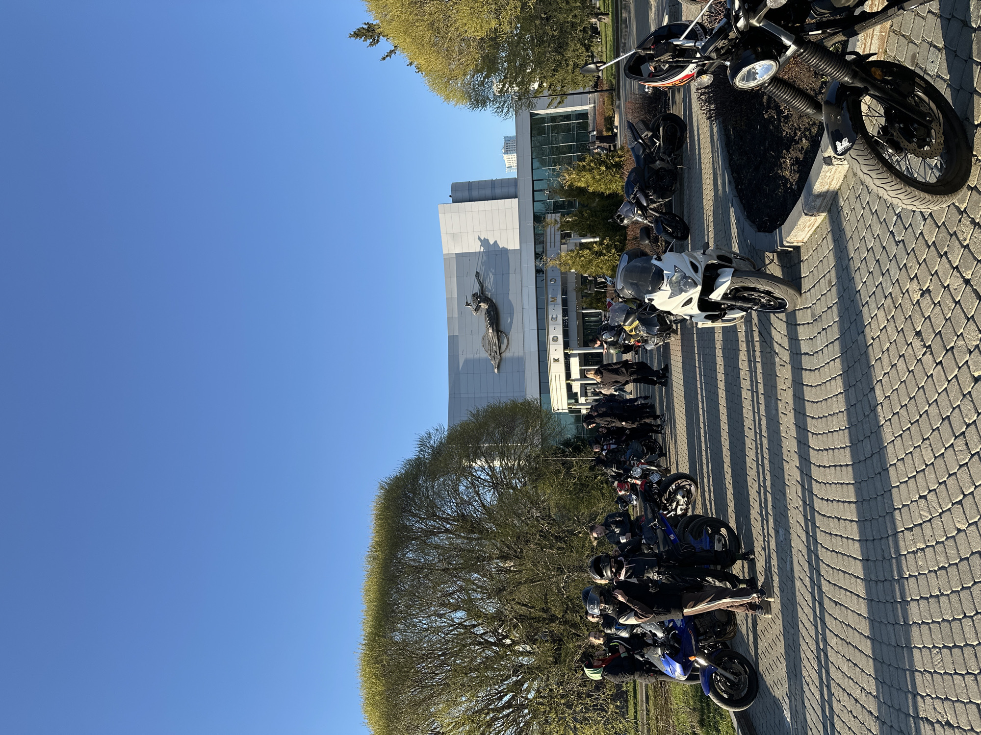The image captures a vibrant outdoor scene on a clear, sunny day, likely during late afternoon or early morning, indicated by the long shadows cast across the paved ground. A large group of people, many dressed in motorcycle gear including helmets and jackets, are gathered around numerous motorcycles of various types, including sport bikes, cruisers, and dual-sports. The motorcycles are parked in a row on a patterned paved area, suggesting a plaza or wide sidewalk.
In the background stands a large, modern building with a light-colored facade. A prominent, dynamic sculpture of a human figure is affixed to the building's exterior, seemingly in mid-air or a flying pose. Below this sculpture and to the right, visible text on the building facade reads "YELTSIN CENTER". This building, along with the surrounding area, appears to be an urban or public space.
To the left of the building, a large tree with light green, newly unfurled leaves signifies springtime. The bright blue sky is clear, indicating pleasant weather suitable for outdoor activities like motorcycling. The overall impression is a social gathering or meet-up of motorcyclists, enjoying the good weather and their shared passion.