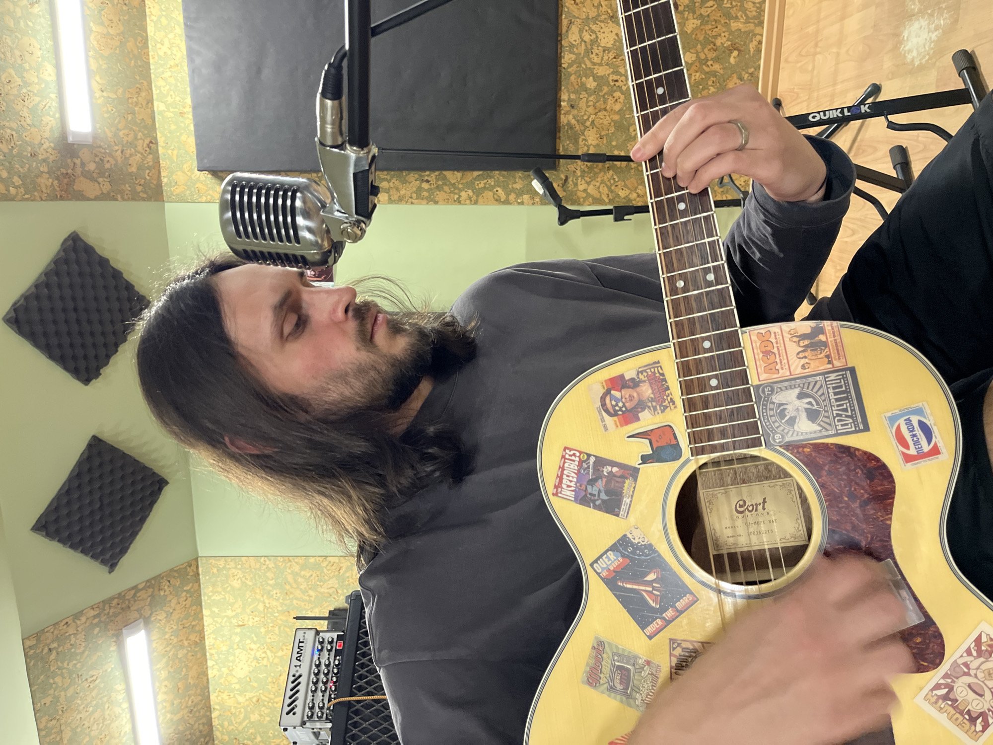 A man with long hair and a beard is playing an acoustic guitar in what appears to be a recording studio. He is positioned in front of a vintage-style microphone on a stand. The guitar is adorned with various stickers featuring bands like AC/DC and Led Zeppelin, as well as movie and soda brand logos. The man's left hand is on the guitar's neck, while his right hand is strumming the strings. The studio setting includes soundproofing panels on the walls, a light fixture, and audio equipment visible in the background. The location is unknown.