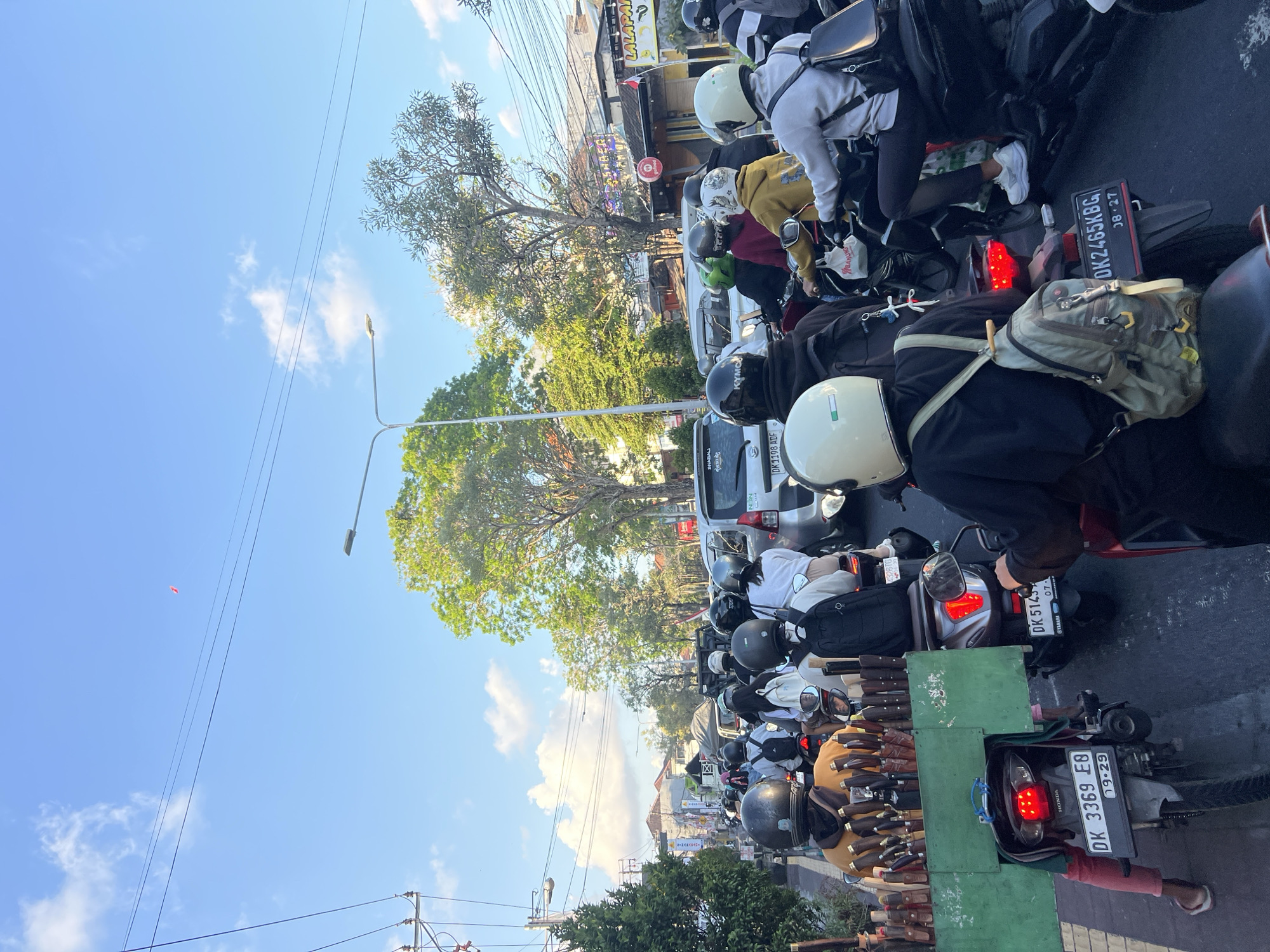 The image depicts a traffic jam in Denpasar, Indonesia, on a sunny day. A multitude of motorbikes and a few cars are stationary on a paved road lined with trees and buildings. People on motorbikes are wearing helmets and backpacks. One person is selling knives from a cart attached to their motorbike. The sky is blue with scattered clouds, and power lines are visible overhead.