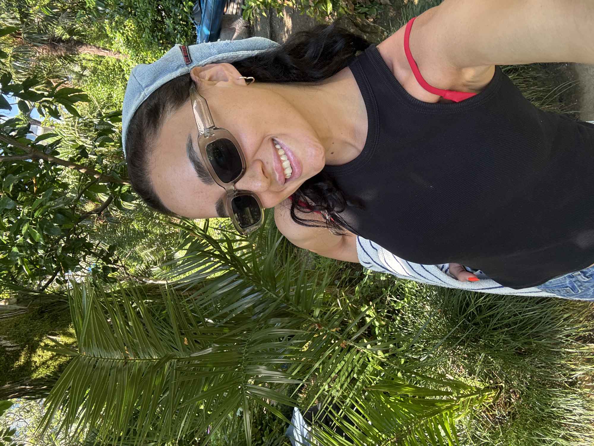 A young woman with dark hair and fair skin is taking a selfie outdoors in a lush, green environment. She is wearing a light blue bandana on her head, dark sunglasses, and a black tank top with a red bra strap visible underneath. A striped garment, possibly a towel or sweater, is draped over her shoulder. Her left arm is extended to take the photo, and she is smiling. The background is filled with vibrant green foliage, including palm fronds and trees, suggesting a tropical or subtropical climate. The lighting indicates it is daytime, with sunlight dappling through the leaves. This scene is consistent with the botanical gardens or a park in Batumi, Georgia, known for its rich flora and subtropical climate.