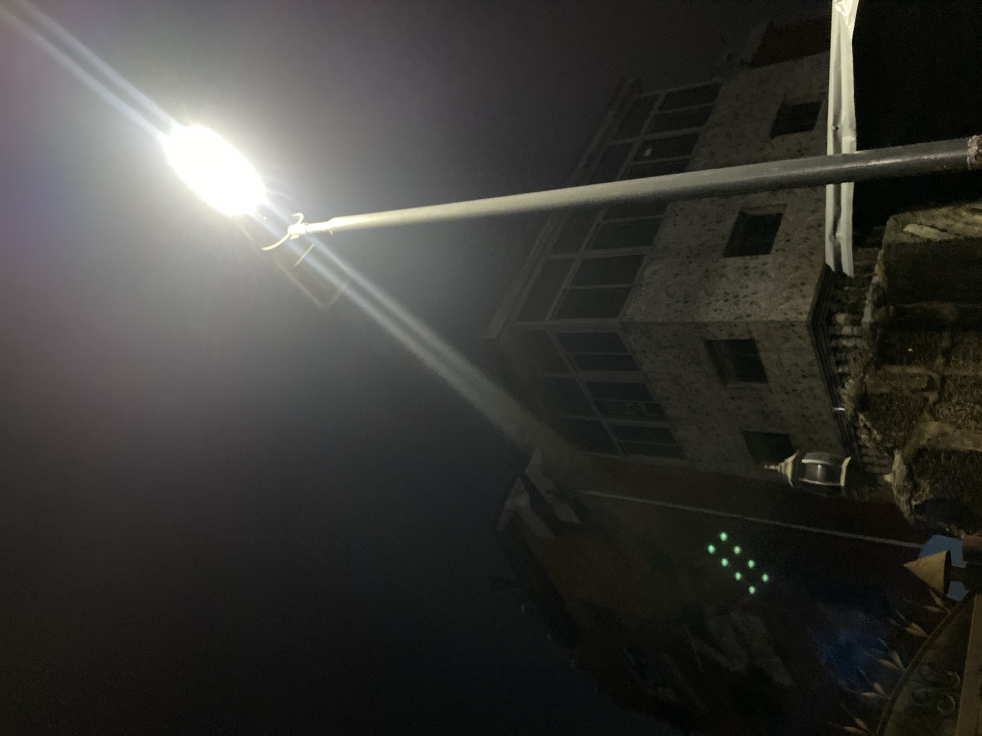 Captured at night, a street lamp, positioned in the upper-left quadrant, radiates a bright light that dominates the frame. The light from the street lamp illuminates a multi-story building located behind and to the right of the light pole. The architecture appears to be modern, featuring regularly spaced windows, suggesting it could be residential or commercial. Adjacent to the building, possibly connected or nearby, is a textured stone or concrete structure. A decorative lamp is also seen. Also, several small green light spots are seen. The surroundings are shrouded in darkness, with only the immediate vicinity illuminated by the street lamp. The image lacks specific contextual clues to definitively place it within Lagos, Nigeria.