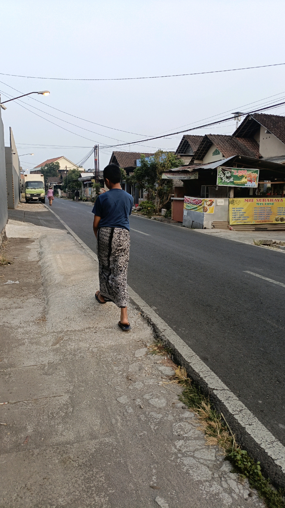 A young person walks along a concrete sidewalk next to a paved road in what appears to be a residential or commercial area of Salatiga, Indonesia. The person is seen from behind, wearing a dark blue t-shirt and a patterned sarong that reaches their ankles, along with dark-colored flip-flops. They are walking away from the camera, towards a row of buildings.
The road is asphalt with a white painted line marking the edge. On the right side of the road, there are several shops or food stalls with awnings and signage. One sign displays "Es Teh 3.000" with an image of iced tea. Another sign, partially visible, seems to advertise "MIE SURABAYA MAS TONO" with a menu of dishes. On the left side of the road, a light green truck is parked. Further down the sidewalk, another person in a pink outfit is visible walking in the same direction as the main subject.
The buildings lining the street have traditional tiled roofs. Overhead, power lines and streetlights are visible against a pale, overcast sky. The sidewalk itself is made of concrete, showing signs of wear and tear with cracks and uneven surfaces. Some dry grass and weeds are growing along the edge of the road where it meets the sidewalk. The overall impression is that of a typical street scene in a town or city in Indonesia during daylight hours.