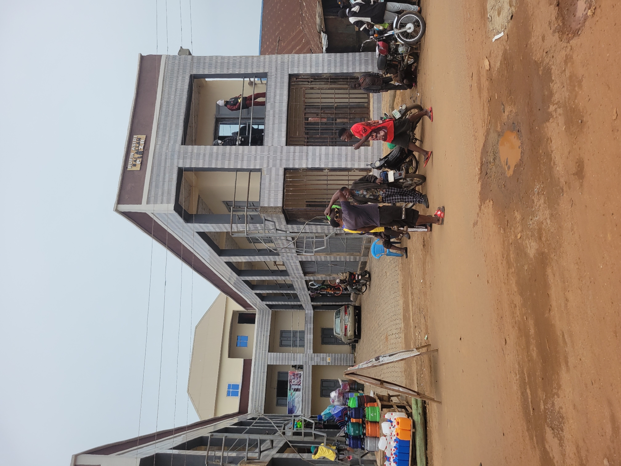 The image depicts an outdoor scene in Bokkos, Nigeria, likely during the daytime. A multi-story building with a modern facade, featuring horizontal gray and white patterned panels, dominates the left side of the frame. A sign on the building reads "RUKIN ASIRI PLAZA".
Several people are present, mostly young men. Some are standing near motorcycles parked on a dirt road in front of the building. One man in a red shirt is leaning towards a motorcycle, appearing to be engaged in conversation or interaction. Another man in a dark shirt and yellow shorts is also near a motorcycle, looking towards the viewer. A person in a red jacket is visible through a window on the second floor.
In the foreground, a stall is set up with various plastic containers and other goods for sale, including colorful tubs and jerrycans. The ground is a dusty, reddish-brown dirt, with some puddles indicating recent rain or dampness. The overall atmosphere seems to be that of a busy street or market area. The lighting suggests it's daytime, with an overcast sky. The image is oriented vertically, with the sky occupying a significant portion of the upper left.