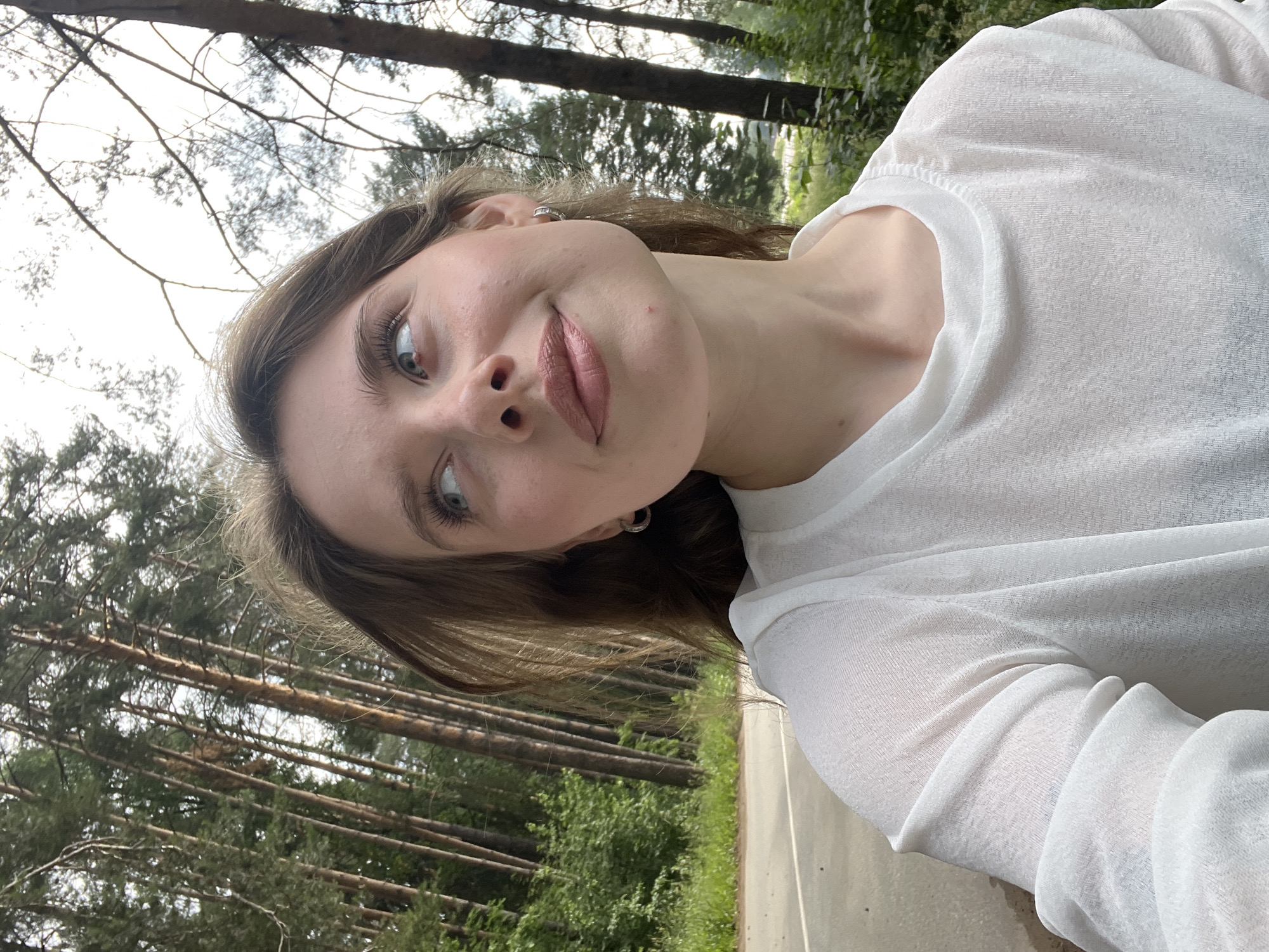 A young woman is taking a selfie outdoors with a forest and a road in the background. The woman has long brown hair and is wearing a white translucent top. She is looking up and to her right with a slight smile. The background consists of tall pine trees and a paved road. The location is the city of Chaikovsky, Russia.