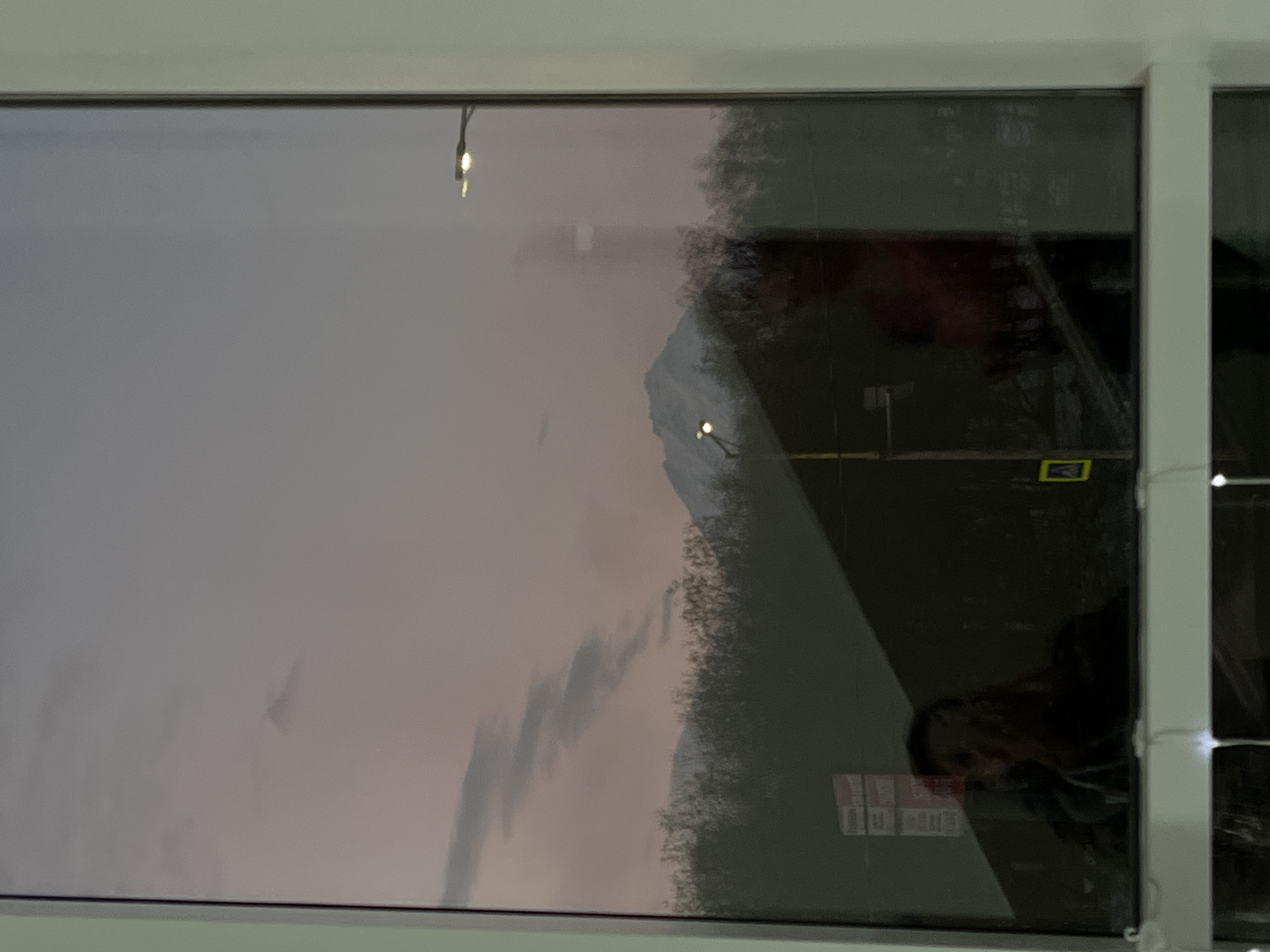 The image depicts a view from a window in Petropavlovsk-Kamchatsky, Russia. The window frame is white, and the glass reflects both the interior and exterior scene. Outside, a muted, perhaps dusk or dawn, sky is visible, along with a snow-capped mountain. Streetlights illuminate the road. A person is reflected in the lower right of the window. Signage is visible, and a pedestrian crossing sign.