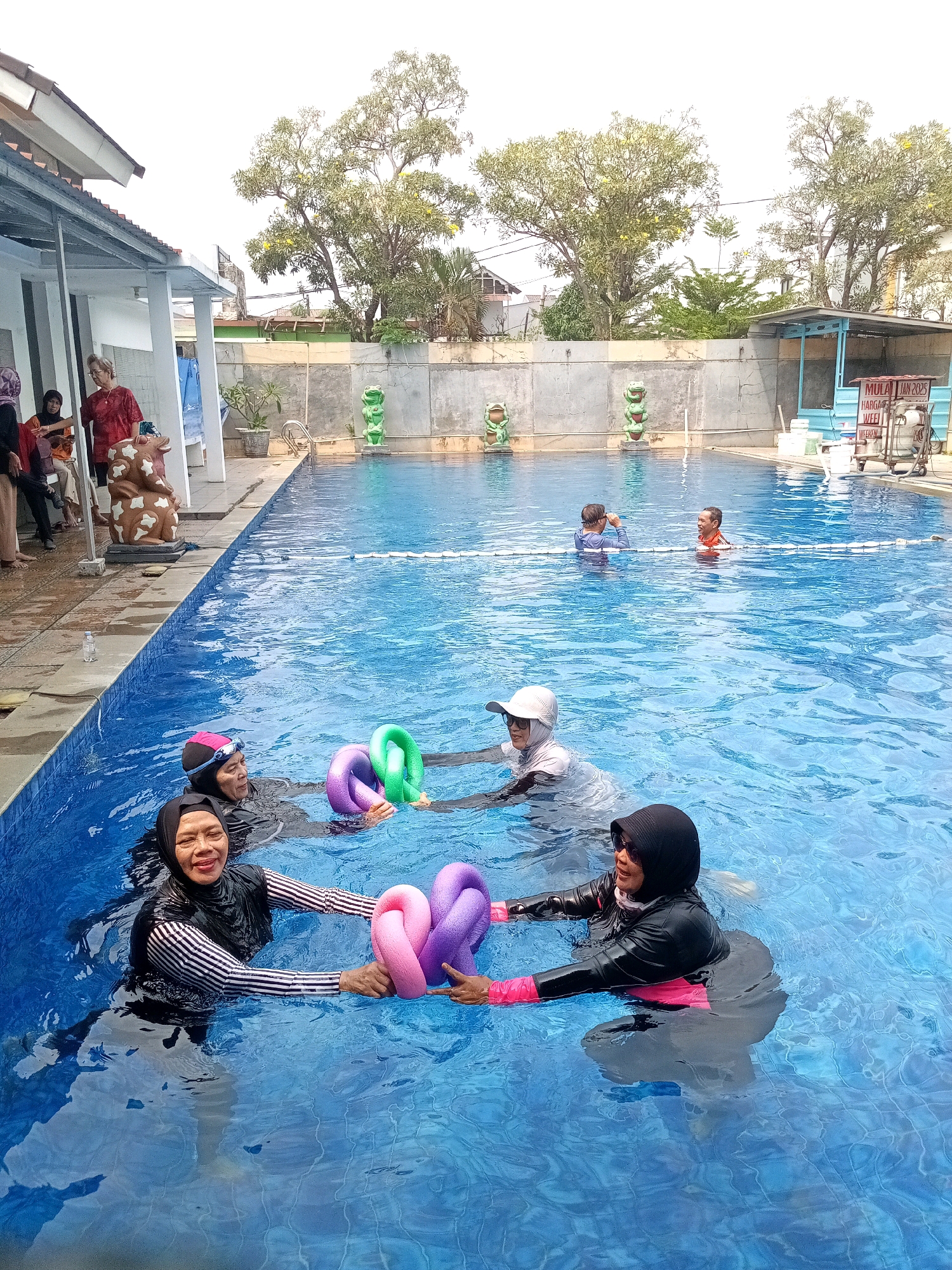 The image depicts an outdoor swimming pool at a recreational facility in Bekasi, Indonesia. In the foreground, four women are in the water, engaged in what appears to be a water aerobics class. Two women are closer to the camera, holding pink and purple pool noodles. One woman, wearing a black and white striped long-sleeved swimsuit and a black hijab, smiles at the camera while holding the noodles. The other woman, in a black swimsuit and black hijab, is looking to the side. Further back in the pool, two more women are visible. One, wearing a white cap and a white rash guard, is looking forward. Another woman, wearing an orange life vest, is visible in the background.
The pool has bright blue water and a tiled edge. To the left of the pool, there is a covered seating area with people sitting and standing. A brown, cow-shaped decorative object is visible near the seating area. In the background, there is a concrete wall with decorative frog statues, and beyond that, trees and buildings, suggesting an urban or suburban environment. A blue sign with Indonesian text is partially visible on the right side of the image. The sky is overcast.