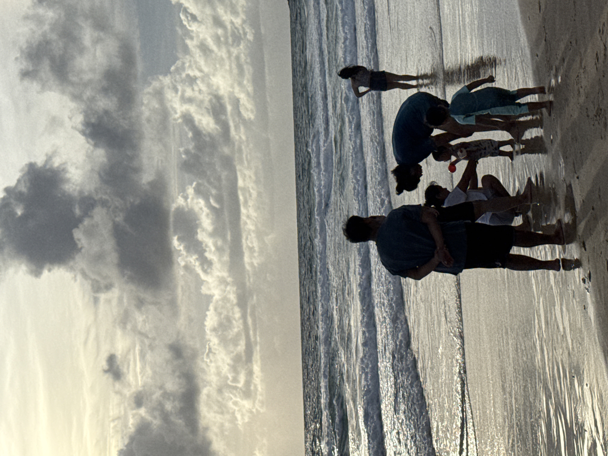 In Karon, Thailand, a family is enjoying a day at the beach. The sun is setting, casting a golden glow over the scene. The ocean waves crash against the shore, and the sky is filled with dramatic clouds. A man and a woman are watching their two children play in the sand. The children are building a sandcastle and splashing in the water. A woman walks in the distance.