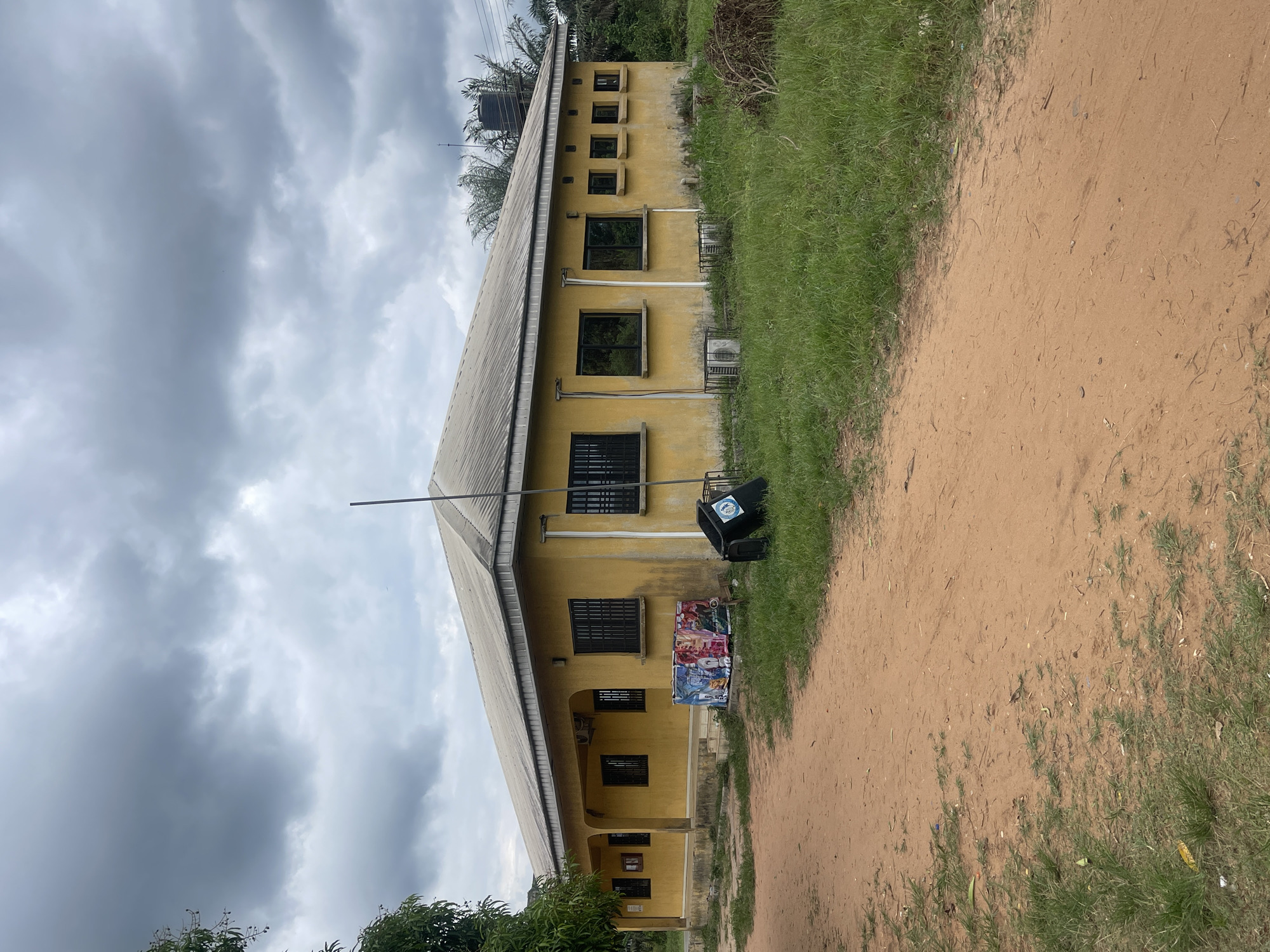 The image shows a two-story yellow building in Abraka, Nigeria, under a cloudy sky. The building has a corrugated metal roof, barred windows, and a tall metal pole extending from the roof. A black water tank is visible on the roof. Beside the building, there is a dirt road with green grass on the edge. A black trash can is positioned near the building, alongside colorful posters or fabrics. Trees and other vegetation are present in the lower left corner of the image. The scene appears to be outdoors in a rural or semi-rural setting.