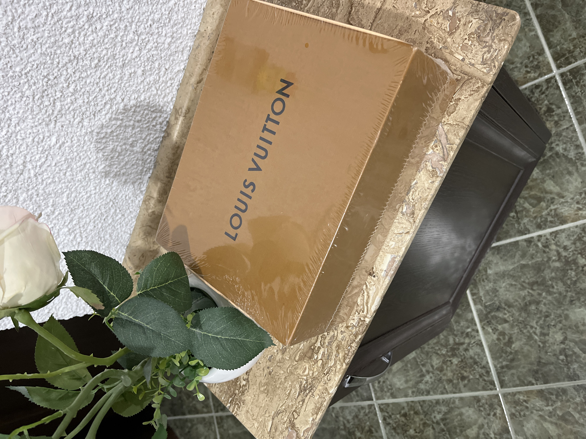 The image depicts a Louis Vuitton box placed on a small, possibly marble-topped, dark brown cabinet. A white rose with green leaves is positioned next to the box on the cabinet. The background includes a white, textured wall and tiled floor. The image does not provide information that could be used to geolocate the scene in Zlatograd, Bulgaria, or anywhere else. No people, actions, or interactions are visible.