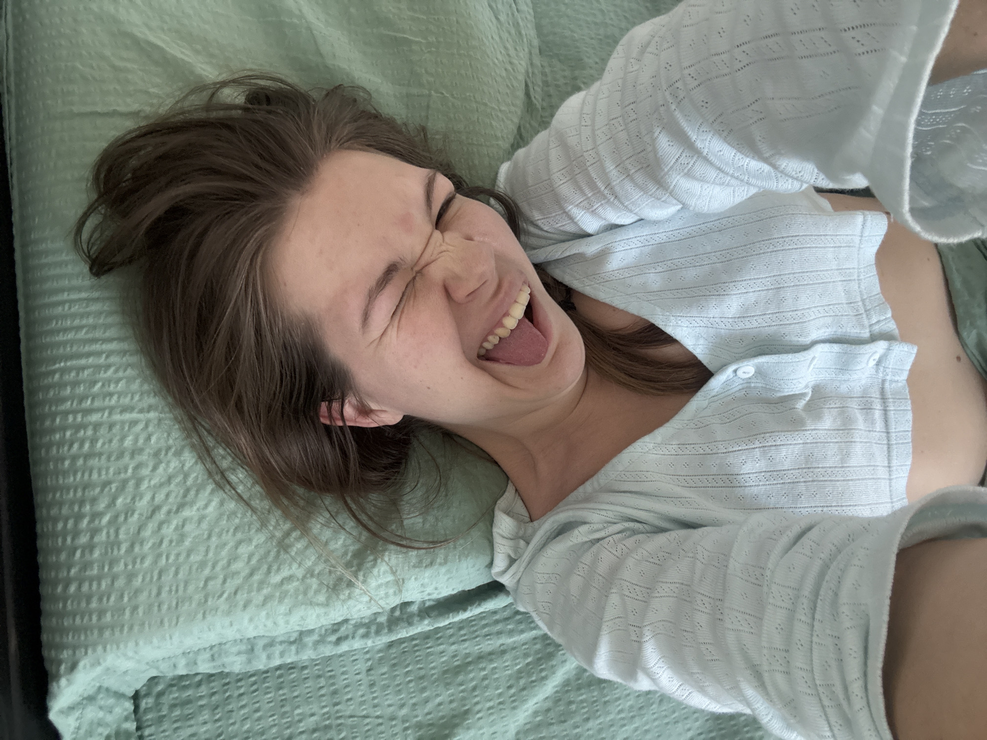 An overhead selfie photograph depicts a single individual, likely female, lying supine on a bed in an indoor setting within Vozdovac Urban Municipality, Serbia. The individual, possessing light brown hair and fair skin, is observed from the mid-chest upwards, with their head positioned towards the bottom of the frame. They are wearing a light blue, long-sleeved, collared knit top featuring a textured, eyelet pattern and a buttoned placket. The individual's eyes are tightly closed, and their mouth is wide open, exposing the tongue and upper and lower teeth, suggesting a moment of intense expression such as laughter or shouting. The bed is covered with a light green, textured bedspread or sheet. The individual's arms are raised above their head, out of frame, consistent with holding a camera for a self-portrait.