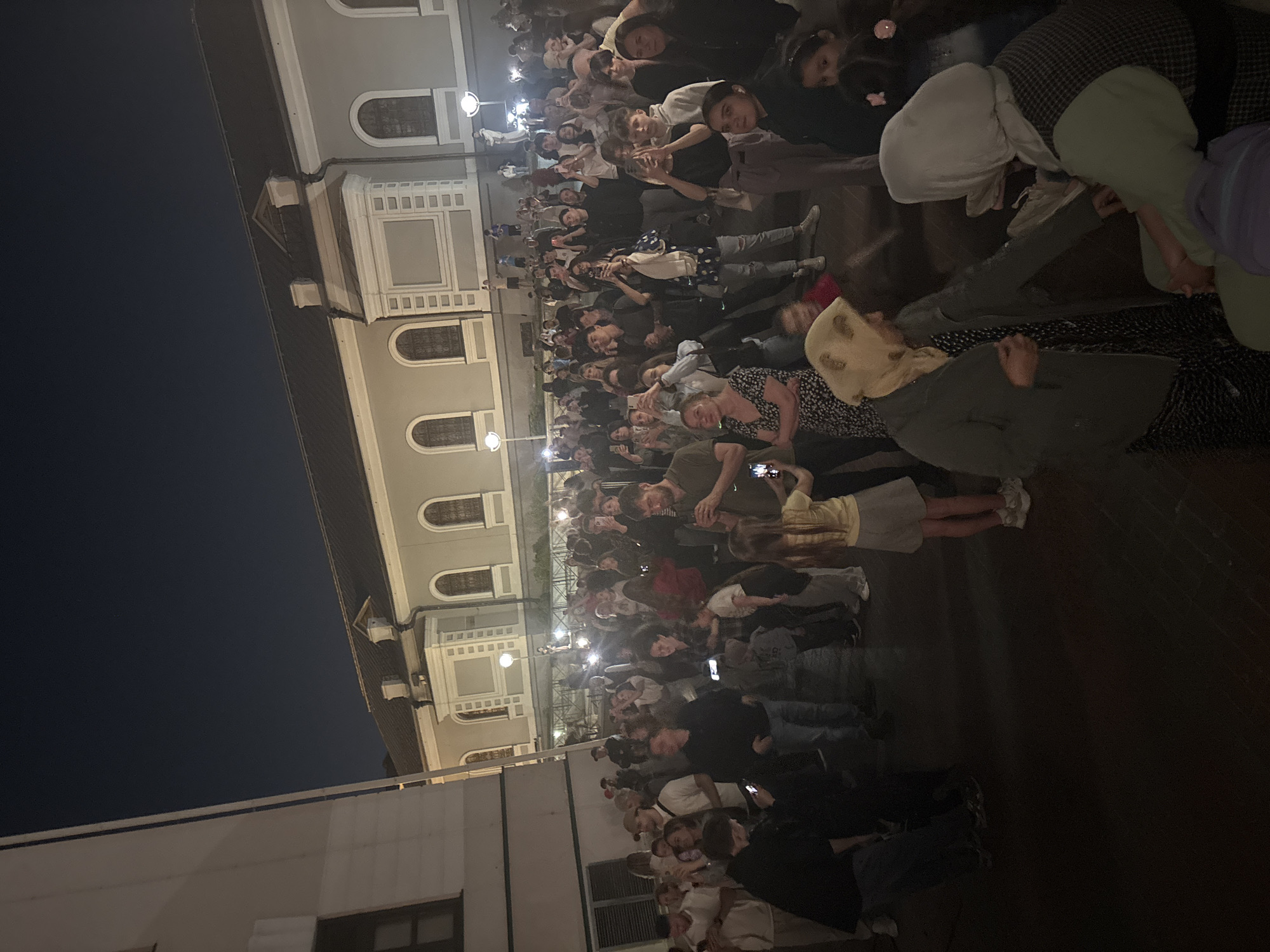 A large group of people stands outside a light gray building in an urban environment at night. Streetlights illuminate the crowd, and some people are looking at and holding their phones, potentially taking pictures or videos. The building has multiple arched windows and architectural details. Some individuals wear head coverings.
