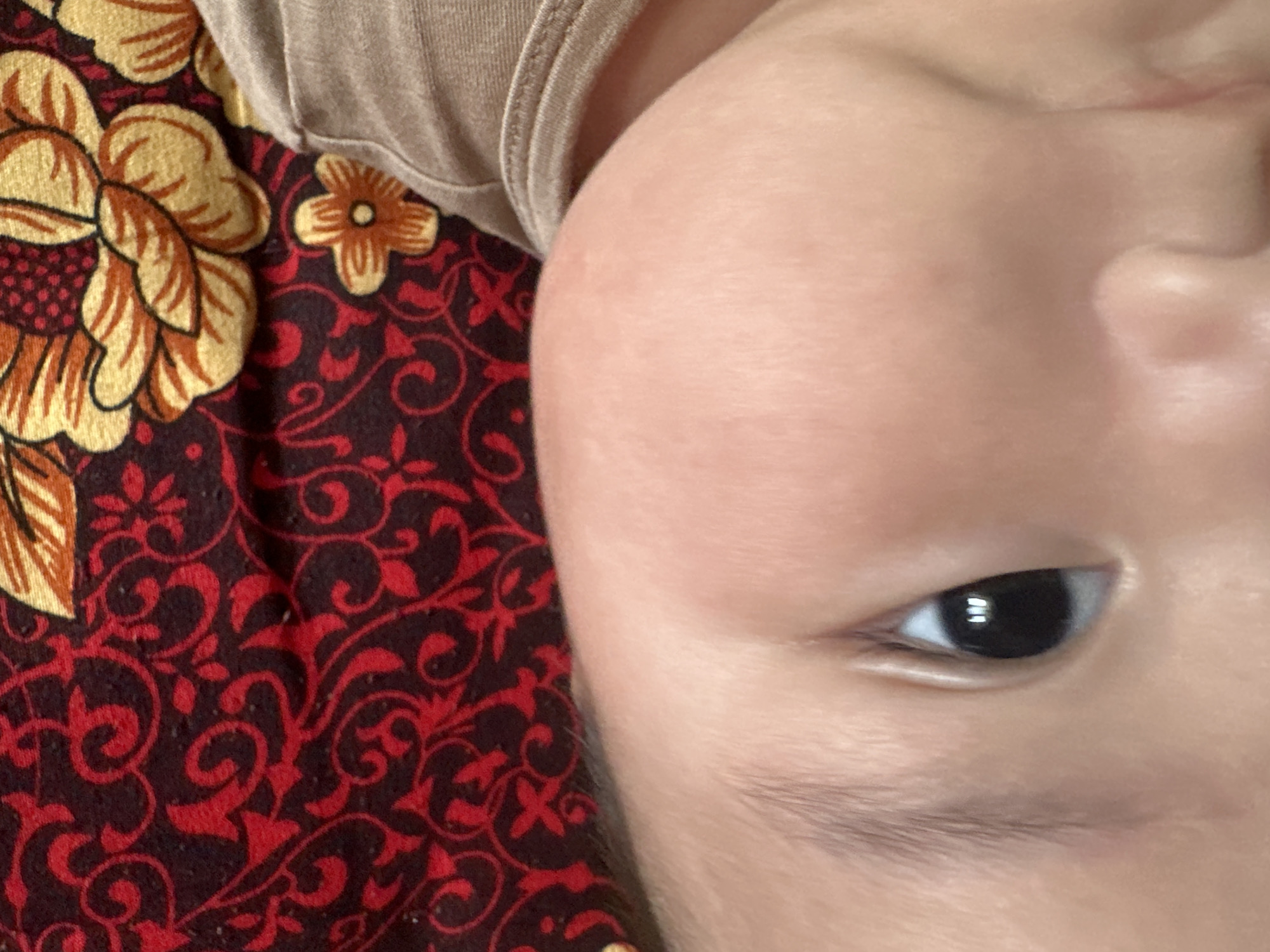 The image is a close-up, rotated view of a baby's face and clothing. The baby is lying on a patterned fabric with a red background and swirling floral designs in red and yellow. The baby's skin is light-toned and smooth, with a visible eye that is dark brown or black. Eyelashes are also visible. Part of the baby's clothing, a beige or light brown fabric with a ribbed texture, is visible at the top of the image. The overall impression is one of a very young child, possibly an infant. The lighting appears to be soft, suggesting an indoor setting. No specific activity or event is depicted, other than the baby resting. There is no visible text in the image. The location context provided is Тайгараев, Kyrgyzstan.