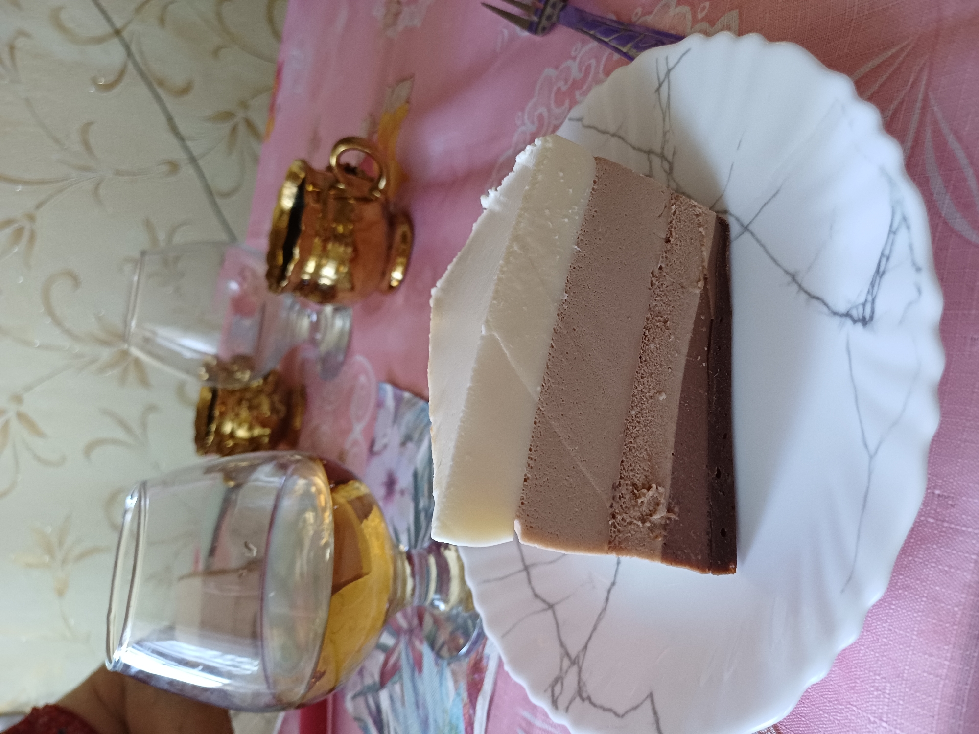 A close-up, eye-level shot shows a slice of three-layered mousse cake on a white, marble-patterned plate. The cake has layers of dark chocolate, milk chocolate, and white chocolate. The plate is set on a pink tablecloth with a lace trim. In the background, two clear glasses and two small golden goblets are visible. One glass is partially filled with a light-colored liquid. A purple fork rests to the right of the cake. The scene is lit by ambient light, casting soft shadows.