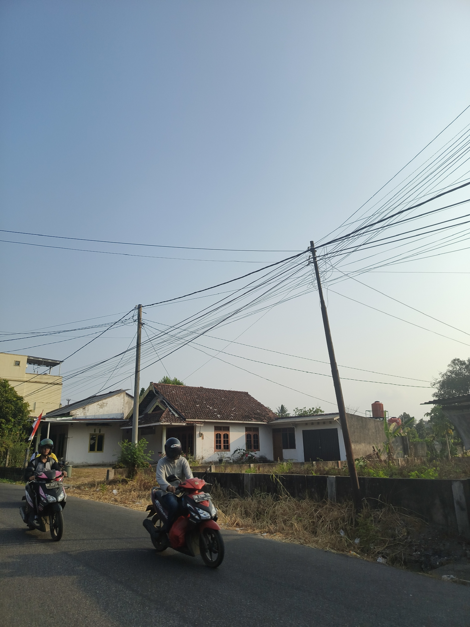 The image depicts a street scene in Pangkalpinang, Indonesia. Two individuals are riding motorcycles on a paved road. The person on the left is riding a pink and black scooter, wearing a yellow shirt and a dark helmet. The person on the right is riding a red and black scooter, wearing a light-colored long-sleeved shirt and a black helmet. Both motorcycles are in motion, heading away from the viewer.
On the right side of the road, there are several houses with tiled roofs and visible windows and doors. One house has a light-colored facade with a red-tiled roof, while another appears to be a more modern structure with a gray concrete wall. Electrical poles with a dense network of overhead wires are prominent throughout the scene, spanning across the sky. Vegetation, including dry grass and some green plants, grows along the side of the road and around the houses. A large orange water tank is visible on top of one of the buildings. The sky is clear and blue, indicating a daytime setting with sunlight casting shadows. A red and white Indonesian flag is mounted on a pole near one of the houses on the left.