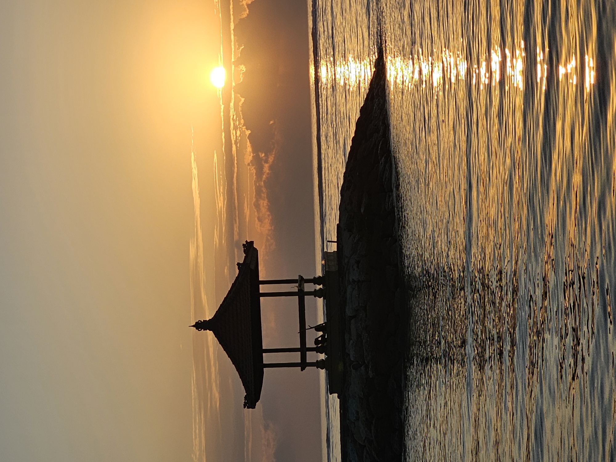 This is a vertical, medium shot photograph of a sunset at Sanur Beach in Denpasar, Indonesia. The sun is a bright, glowing orb, partially obscured by scattered clouds. Its light casts a warm, golden hue across the sky and water, creating a path of shimmering reflections on the ocean's surface. In the foreground, a traditional Balinese gazebo, silhouetted against the vibrant sky, stands on a rocky outcrop. The gazebo has an ornate, tiered roof and is supported by pillars. Inside the gazebo, two figures are visible, appearing as dark silhouettes, one seated and the other possibly leaning or standing. The waves gently lap against the rocks and the shore, and the overall atmosphere is serene and picturesque, typical of a beach sunset in Bali.